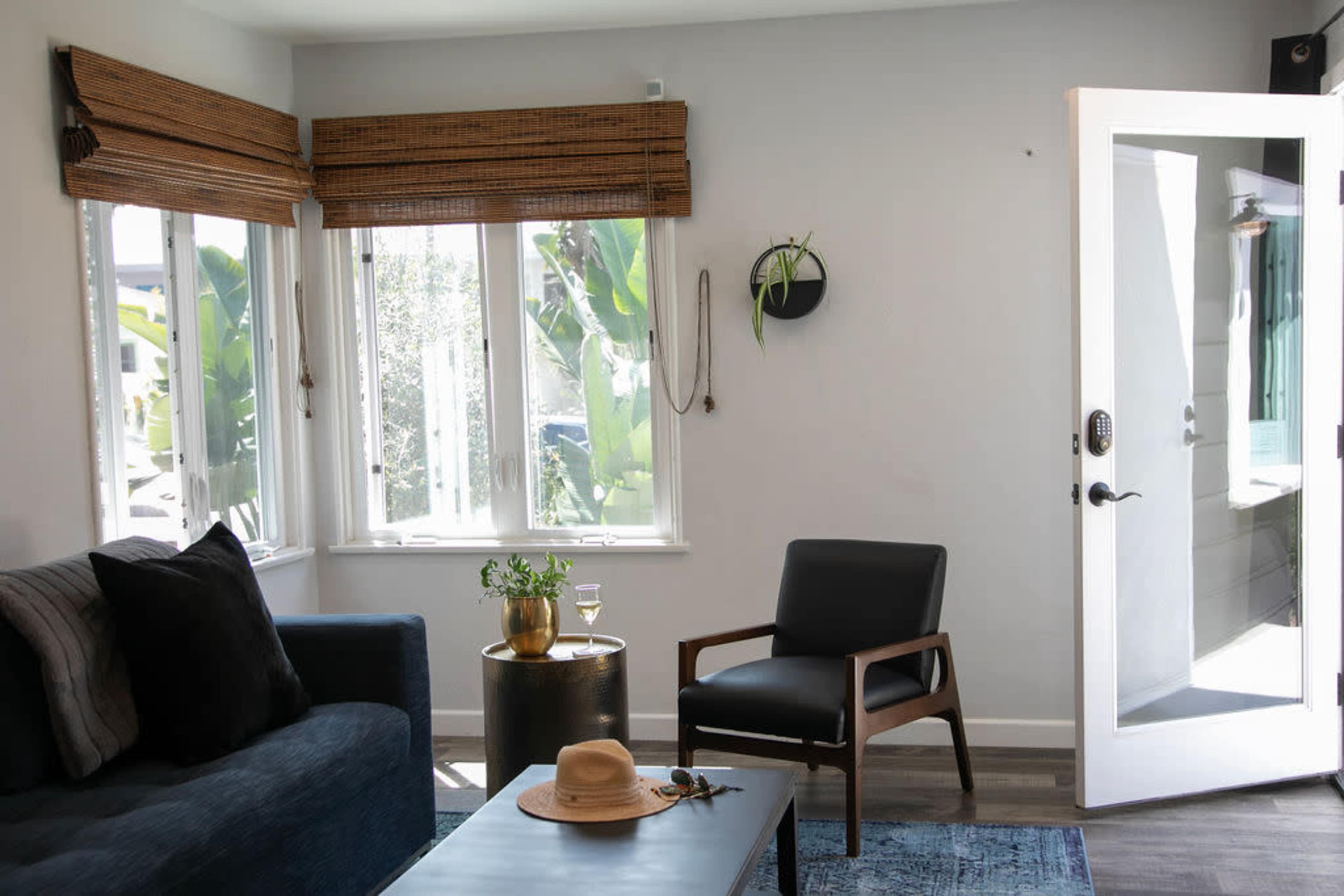 A living room features a dark sofa, a single black chair, a small side table with a drink, and large windows allowing natural light to enter.