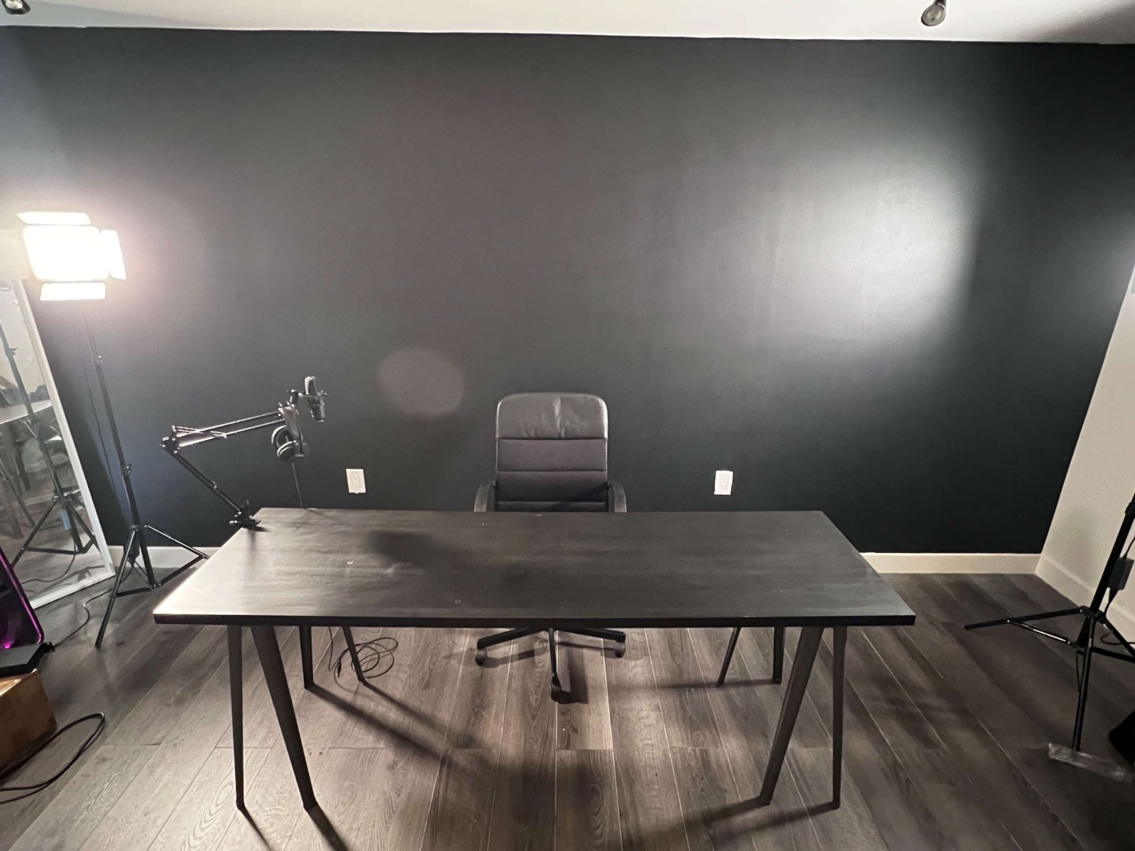A minimalist office setup features a black desk and chair against a dark wall, with soft lighting and a microphone attached to a stand nearby.