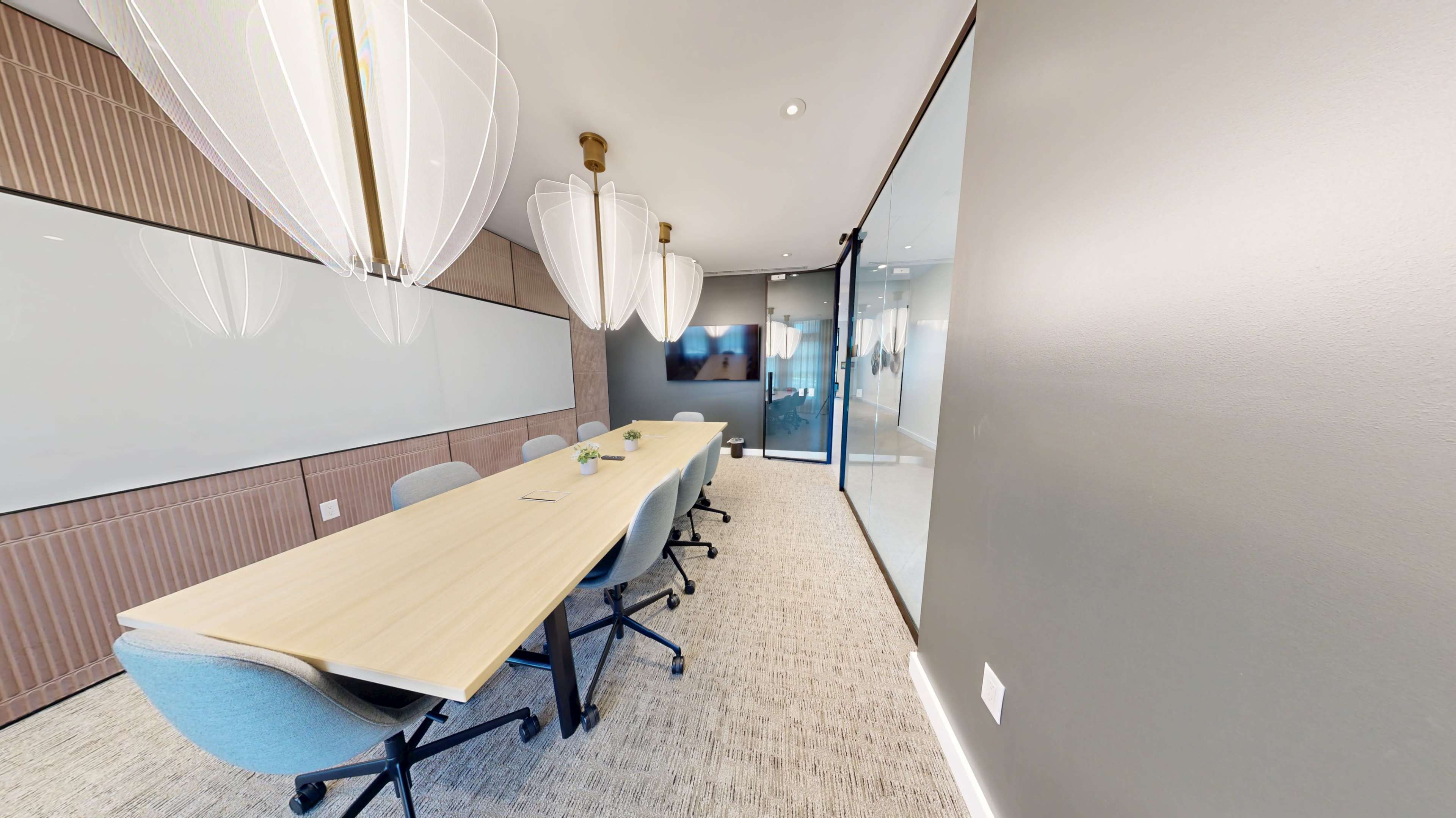The image shows a modern conference room featuring a long wooden table surrounded by office chairs, with large pendant lights hanging overhead and a glass wall on one side.