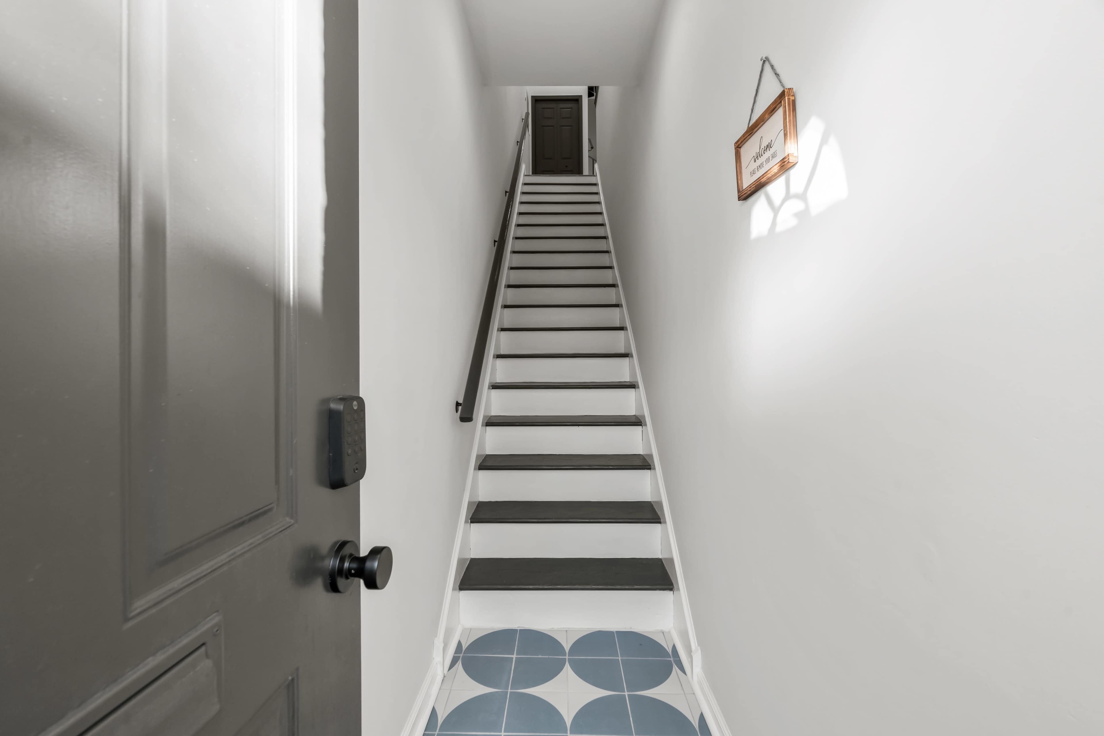 A narrow hallway leads to a staircase with white risers and dark treads, featuring a decorative tile floor at the bottom.