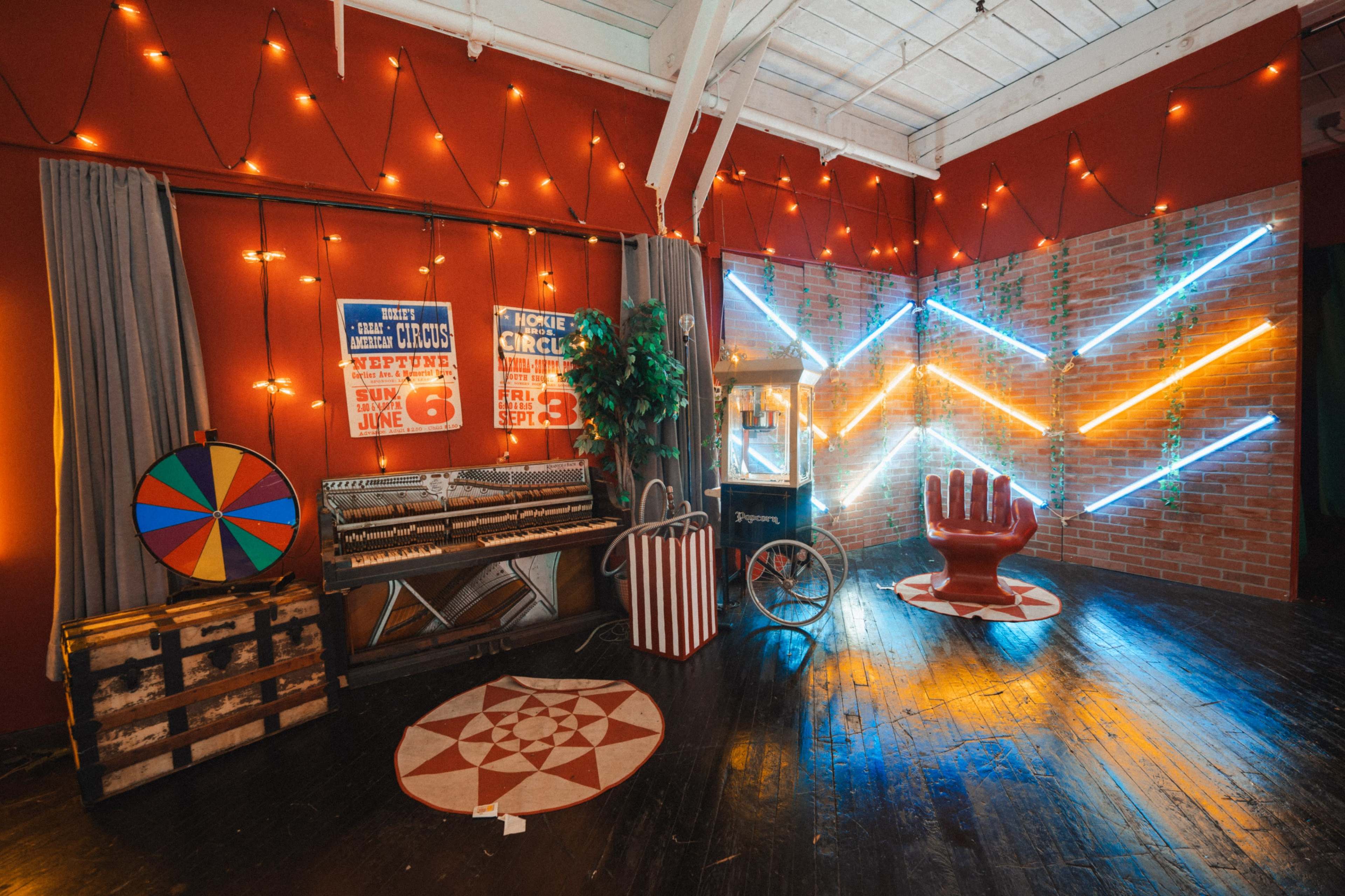 The image shows a vintage-themed room decorated with carnival posters, colorful string lights, a classic popcorn machine, a red hand-shaped chair, and a game wheel.