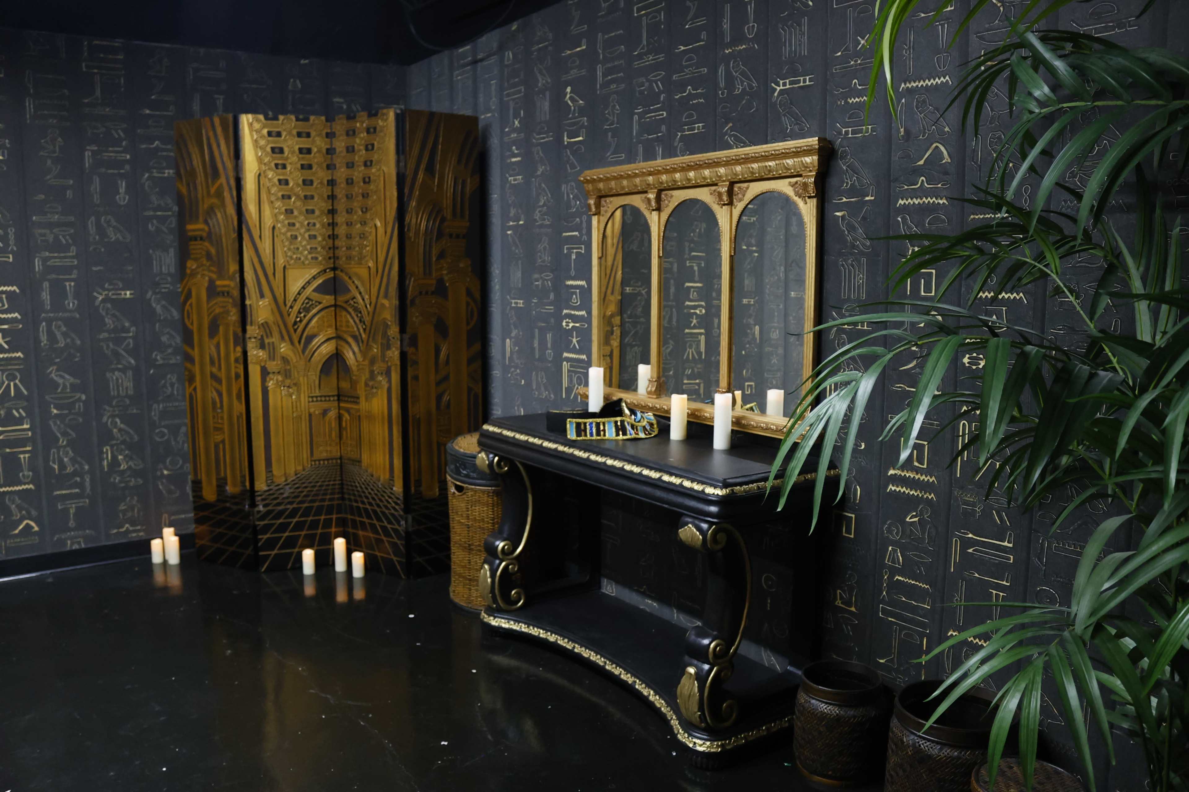 A room with a black wall featuring hieroglyphics, a decorative golden folding screen, a dark console table with a large mirror, and several lit candles.