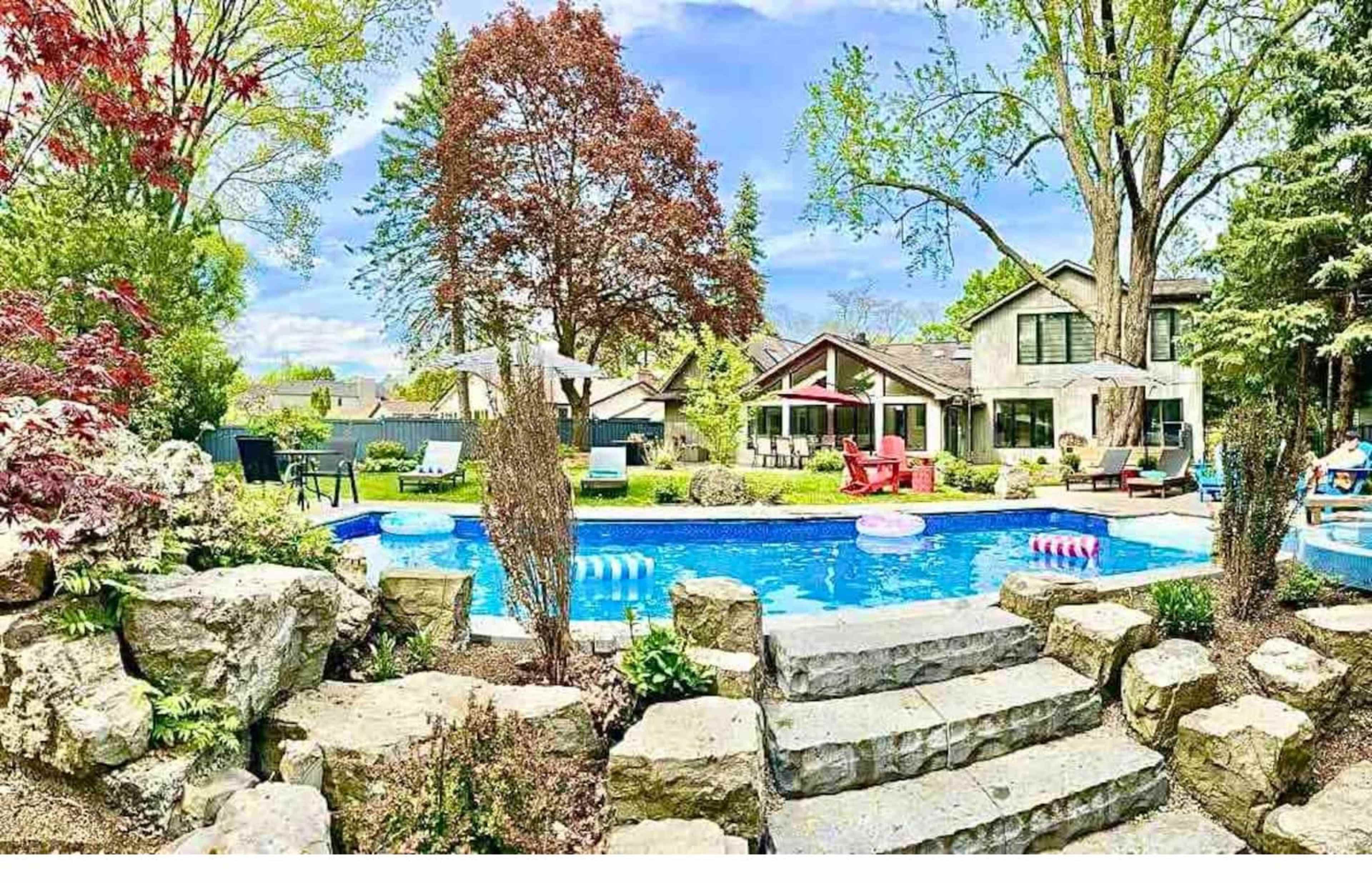 A landscaped backyard features a blue swimming pool surrounded by stone steps, colorful lounge chairs, and lush greenery.