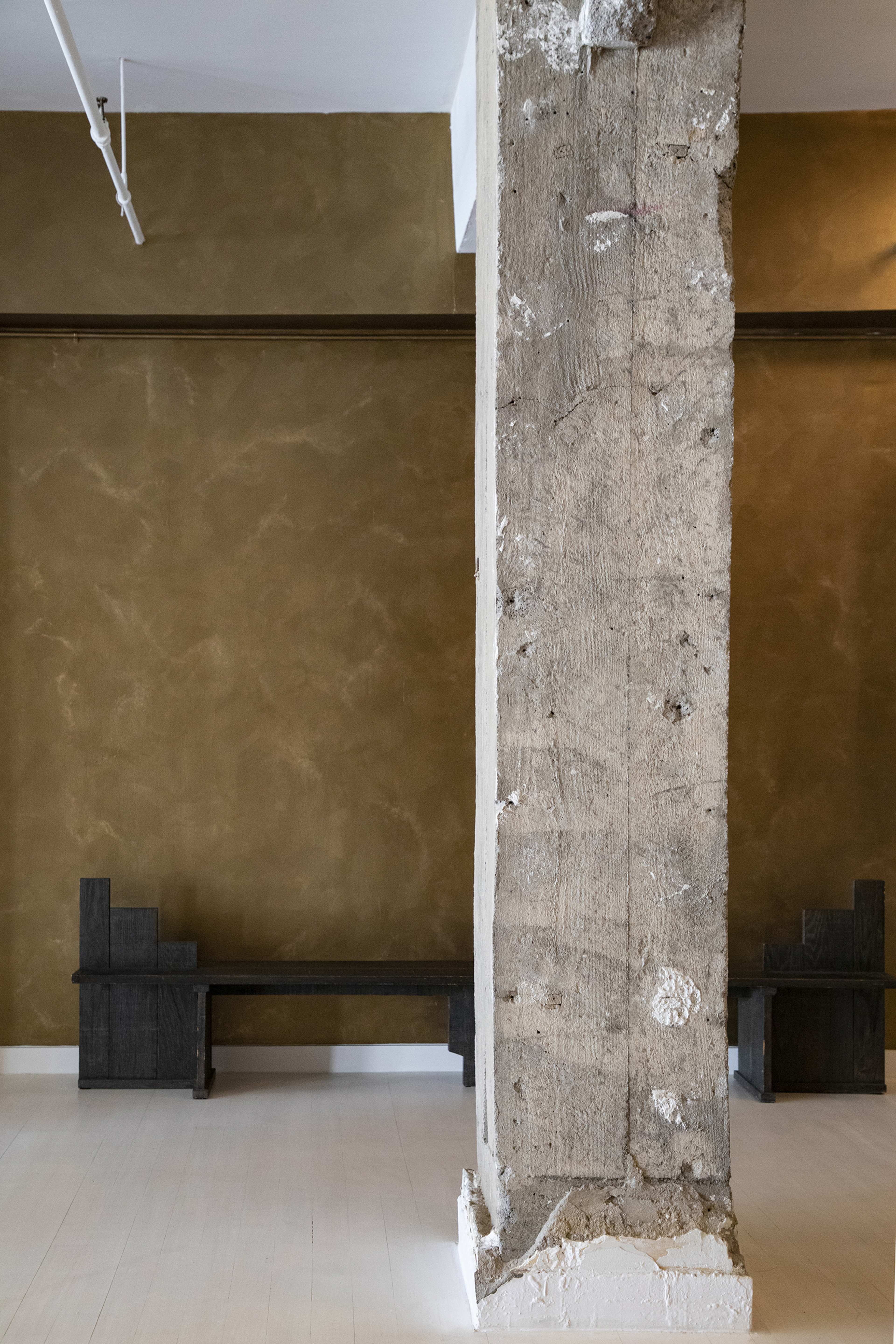 A minimalist interior with a textured brown wall, a concrete pillar in the foreground, and dark wooden benches positioned against the wall.