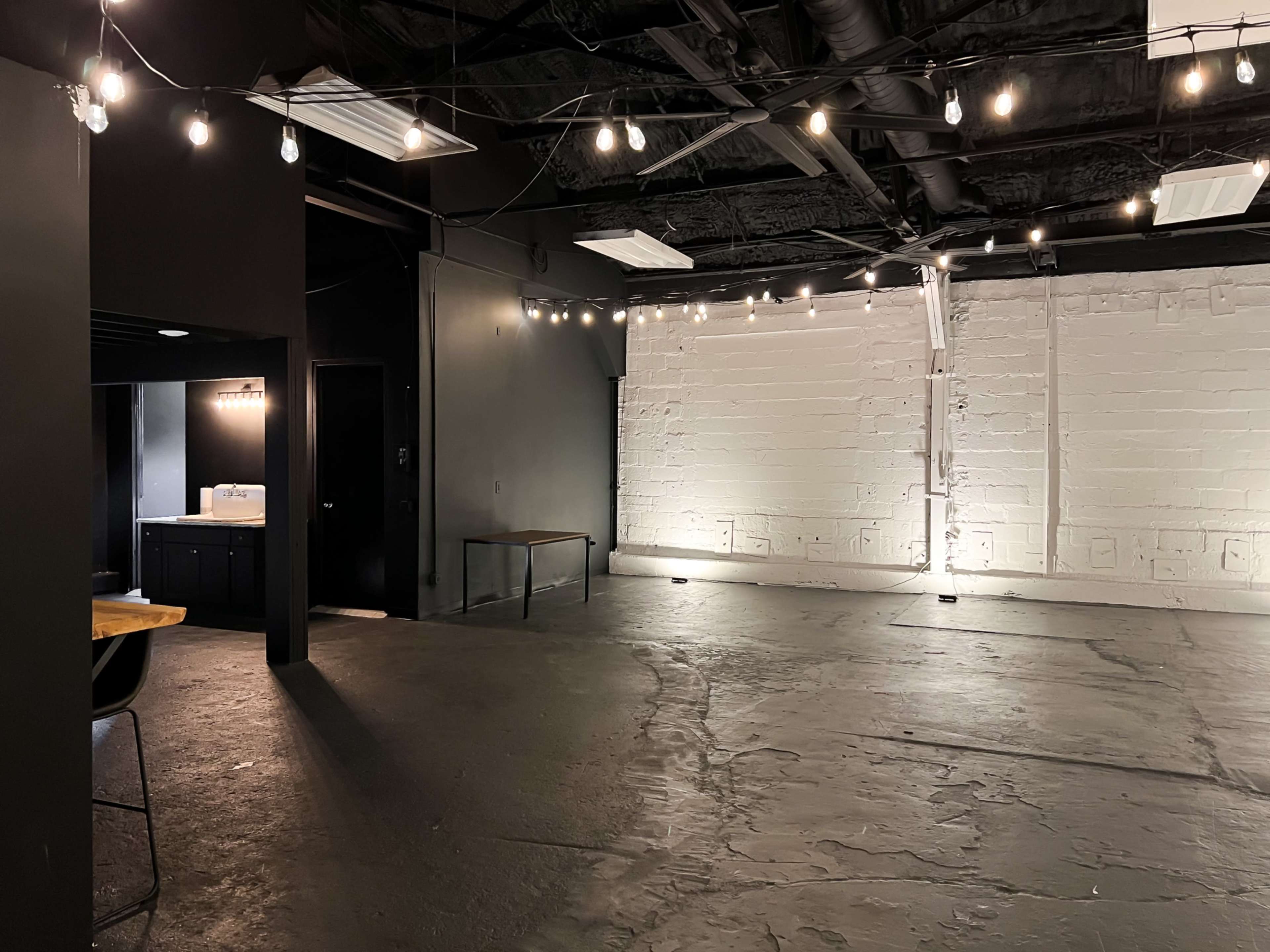 A spacious, dimly lit room with exposed black walls, a concrete floor, and string lights overhead, featuring a small kitchenette area and a table in one corner.