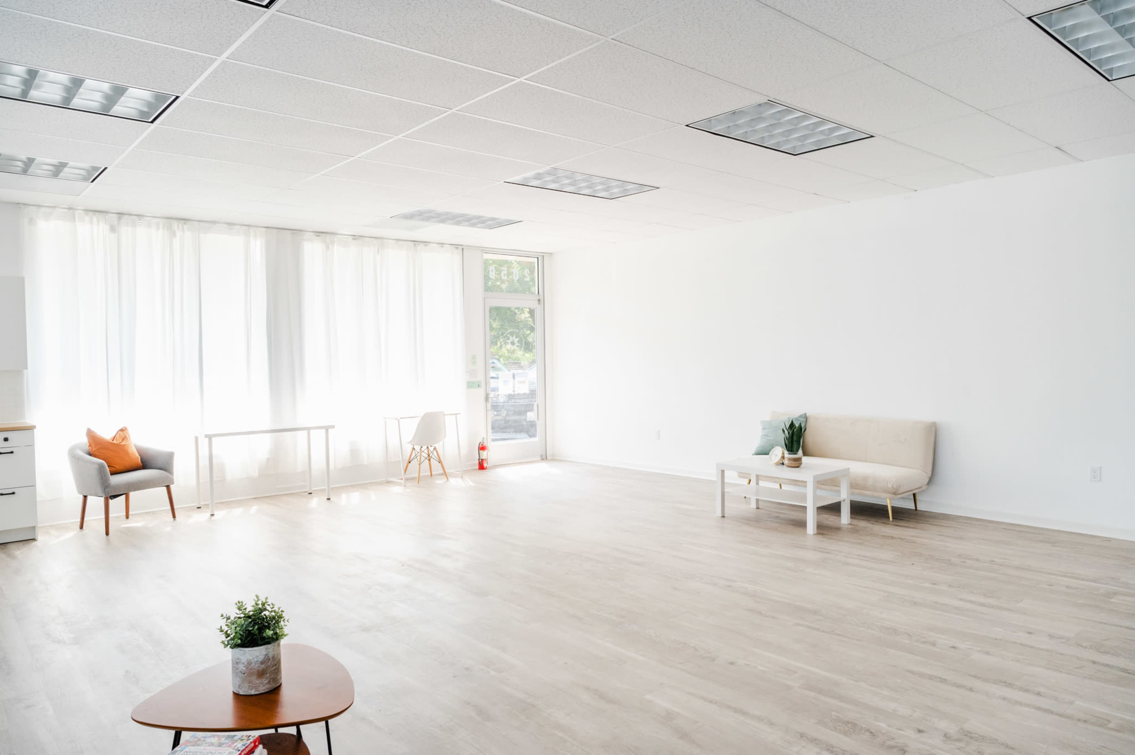 The image shows a spacious, minimalist room with light-colored wooden flooring, large windows, a sofa, a coffee table, and a small desk with a chair.