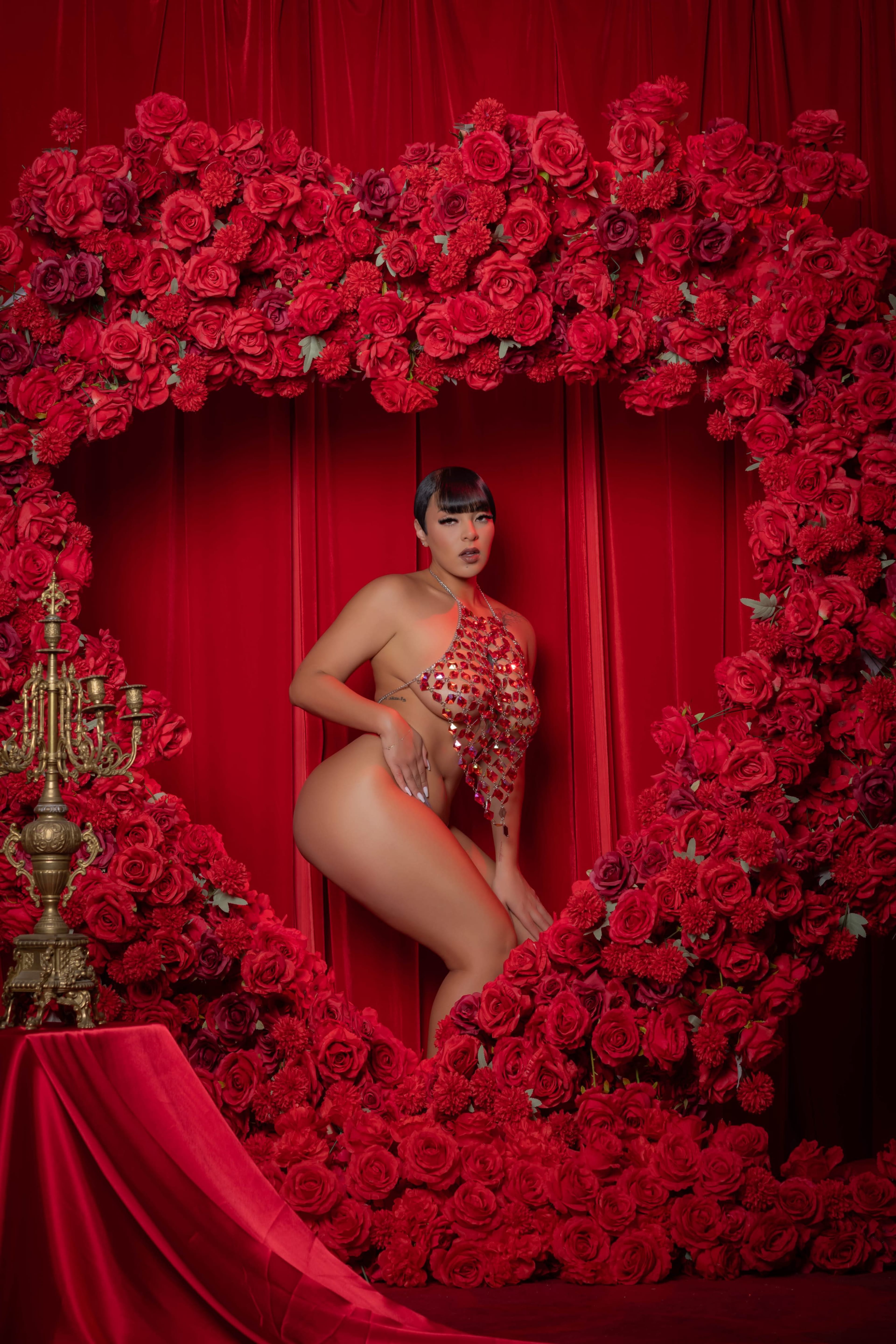 A person poses in front of a large heart-shaped arrangement made of red roses, with red fabric draping the background.