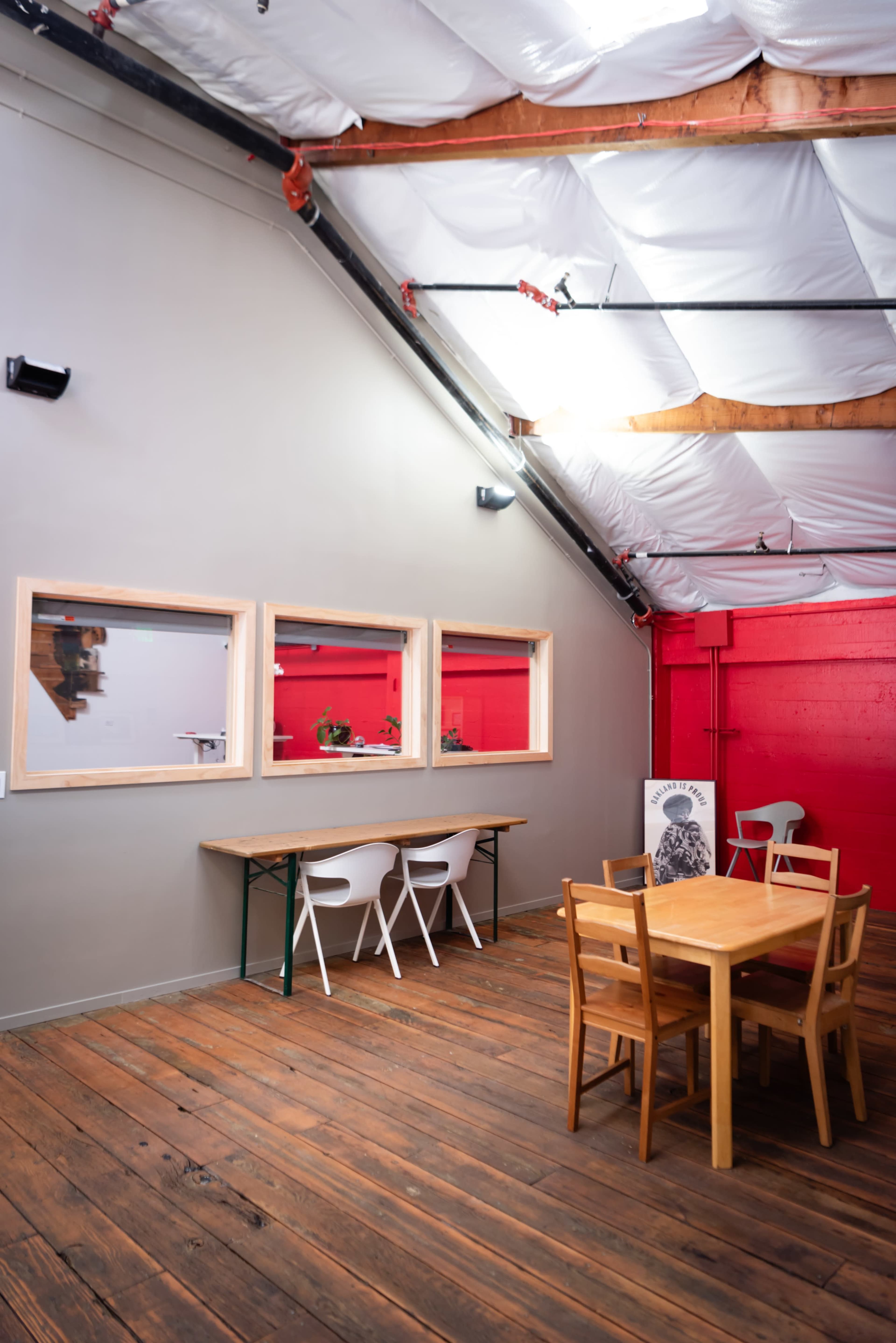The image shows a spacious interior with wooden flooring, featuring a table and chairs in the foreground and large windows along one wall.