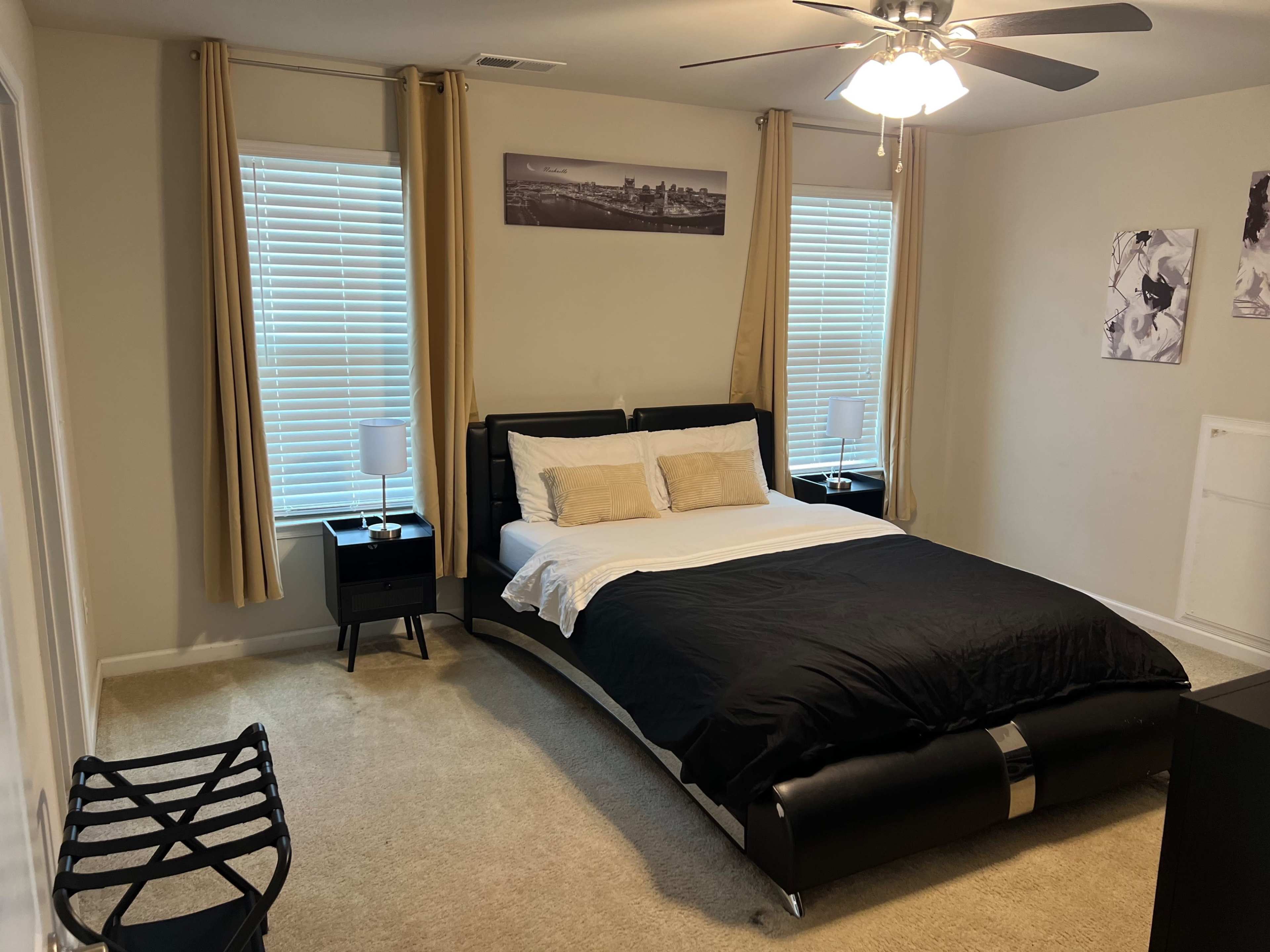 The room features a bed with black bedding, two windows with curtains, nightstands on each side, and a light-colored carpet.