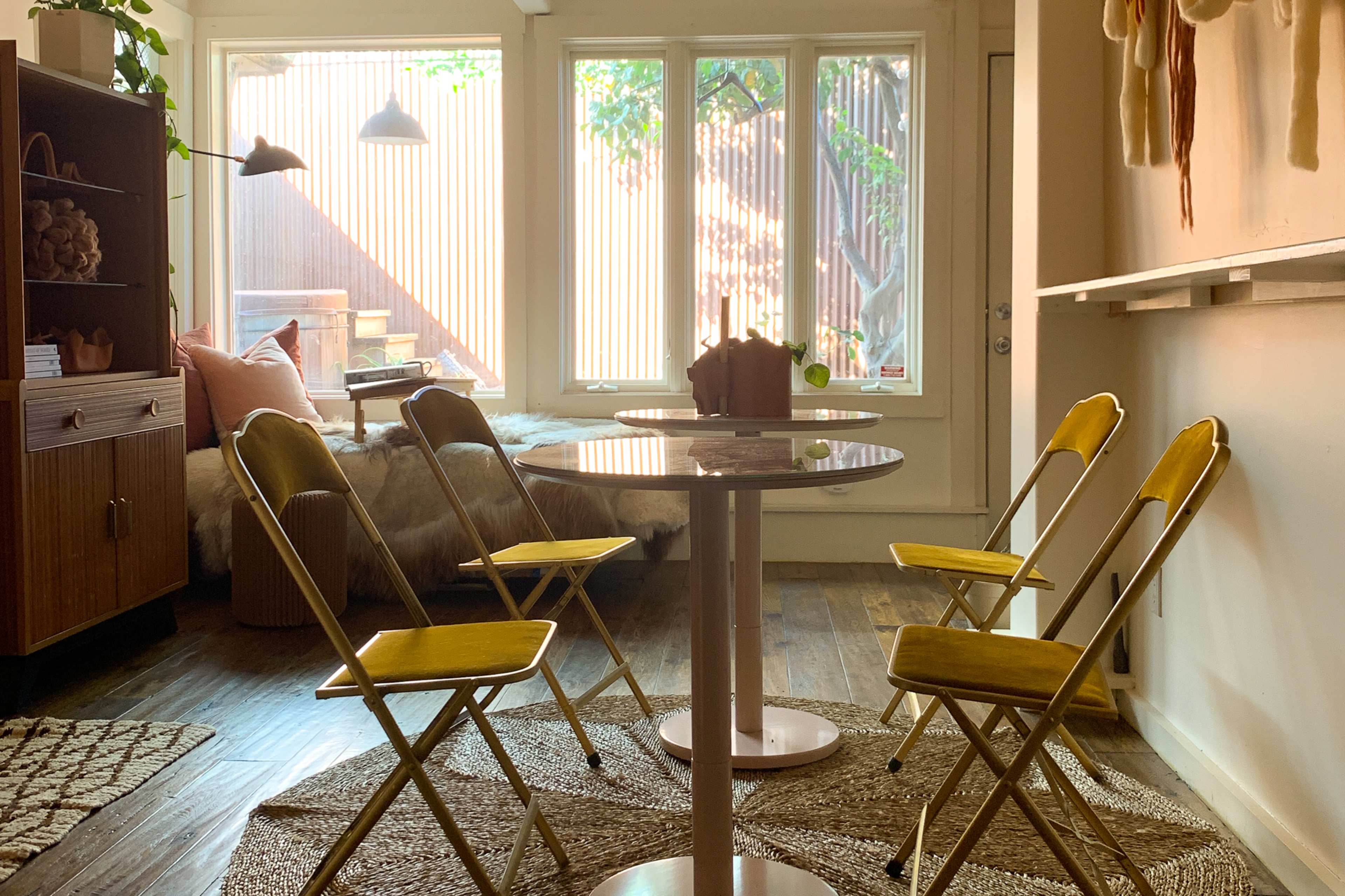 A cozy room with a circular table surrounded by four yellow folding chairs, soft seating, and large windows letting in natural light.