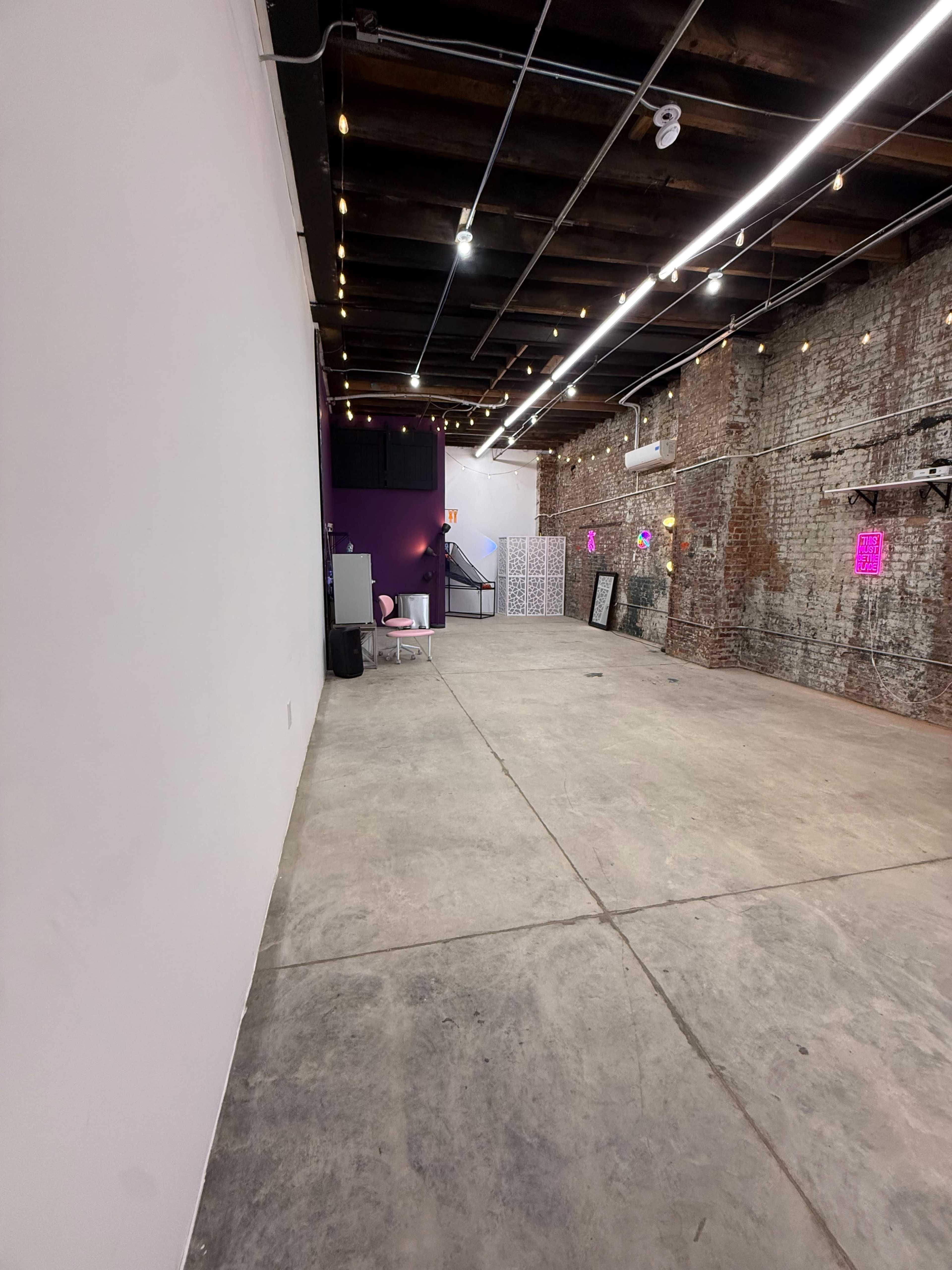 The image shows a spacious interior with exposed brick walls, a concrete floor, and overhead string lights, featuring a staircase at the far end.