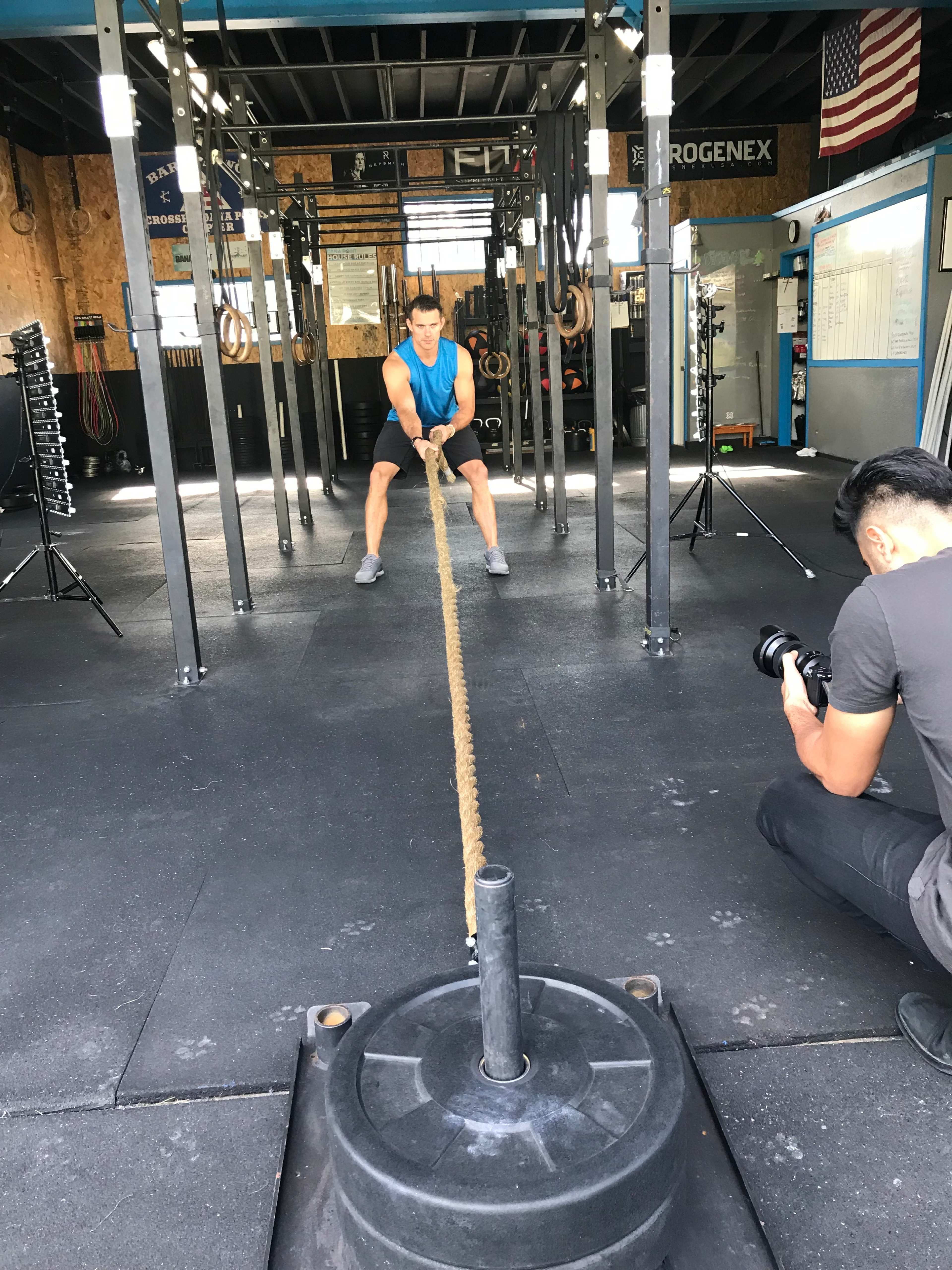A person is pulling a heavy rope attached to a large weight in a gym while another individual records the activity with a camera.