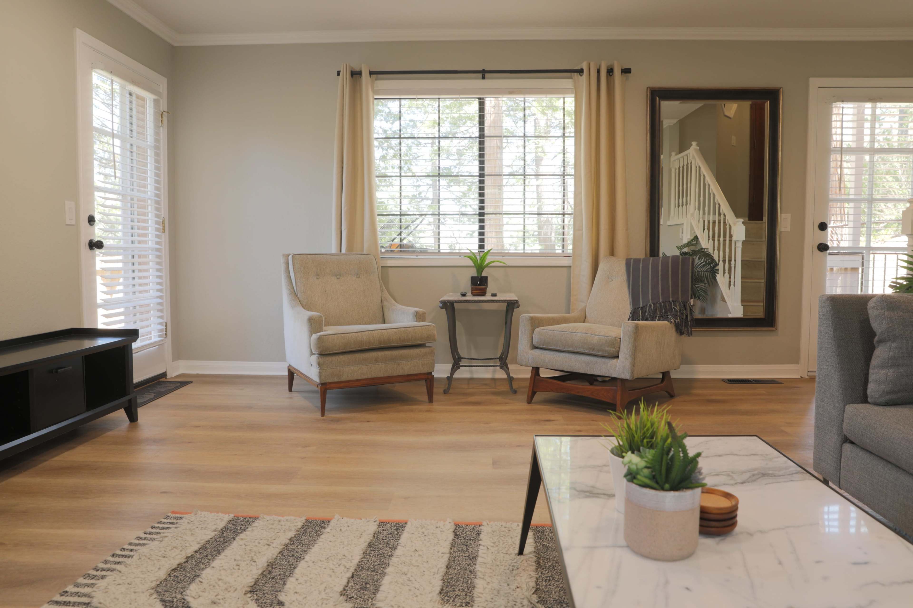 The living room features two armchairs, a small side table, and a large mirror, with natural light streaming through a window.
