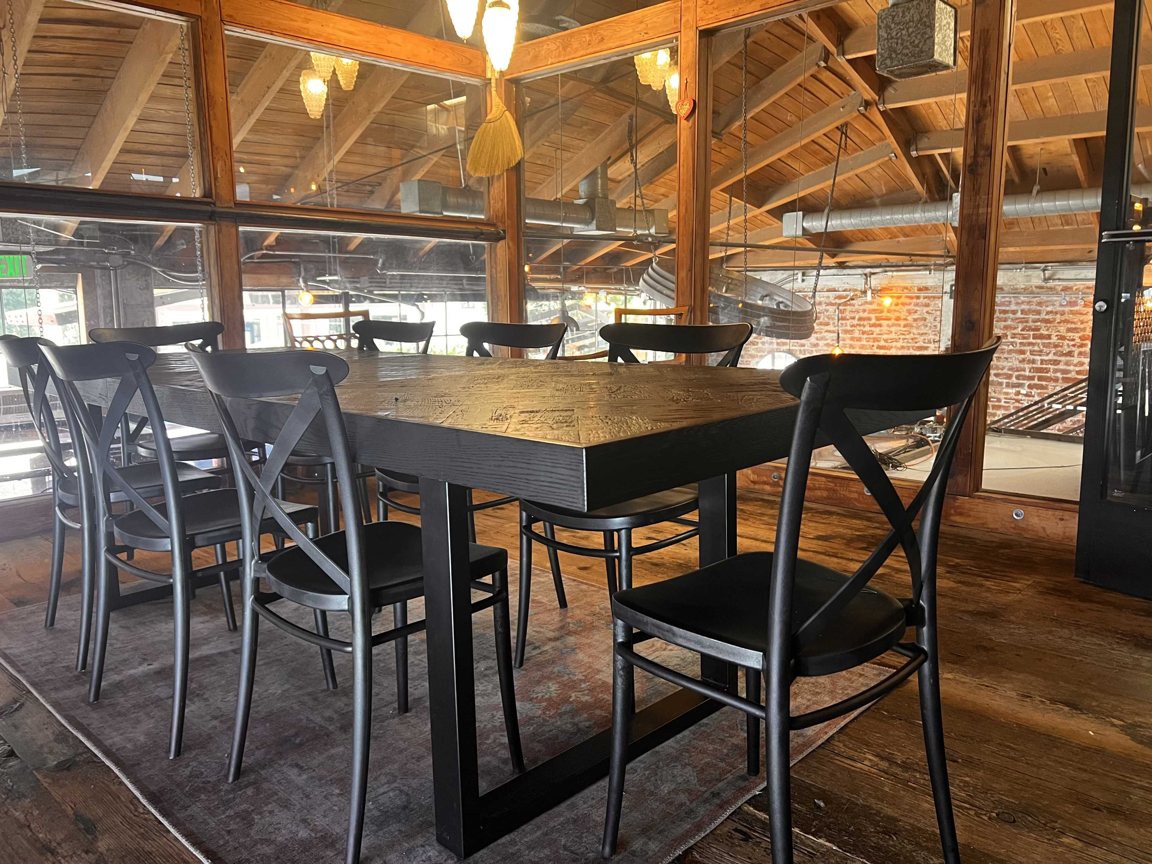 A large wooden dining table surrounded by black chairs is set in a room with exposed brick walls and large windows.