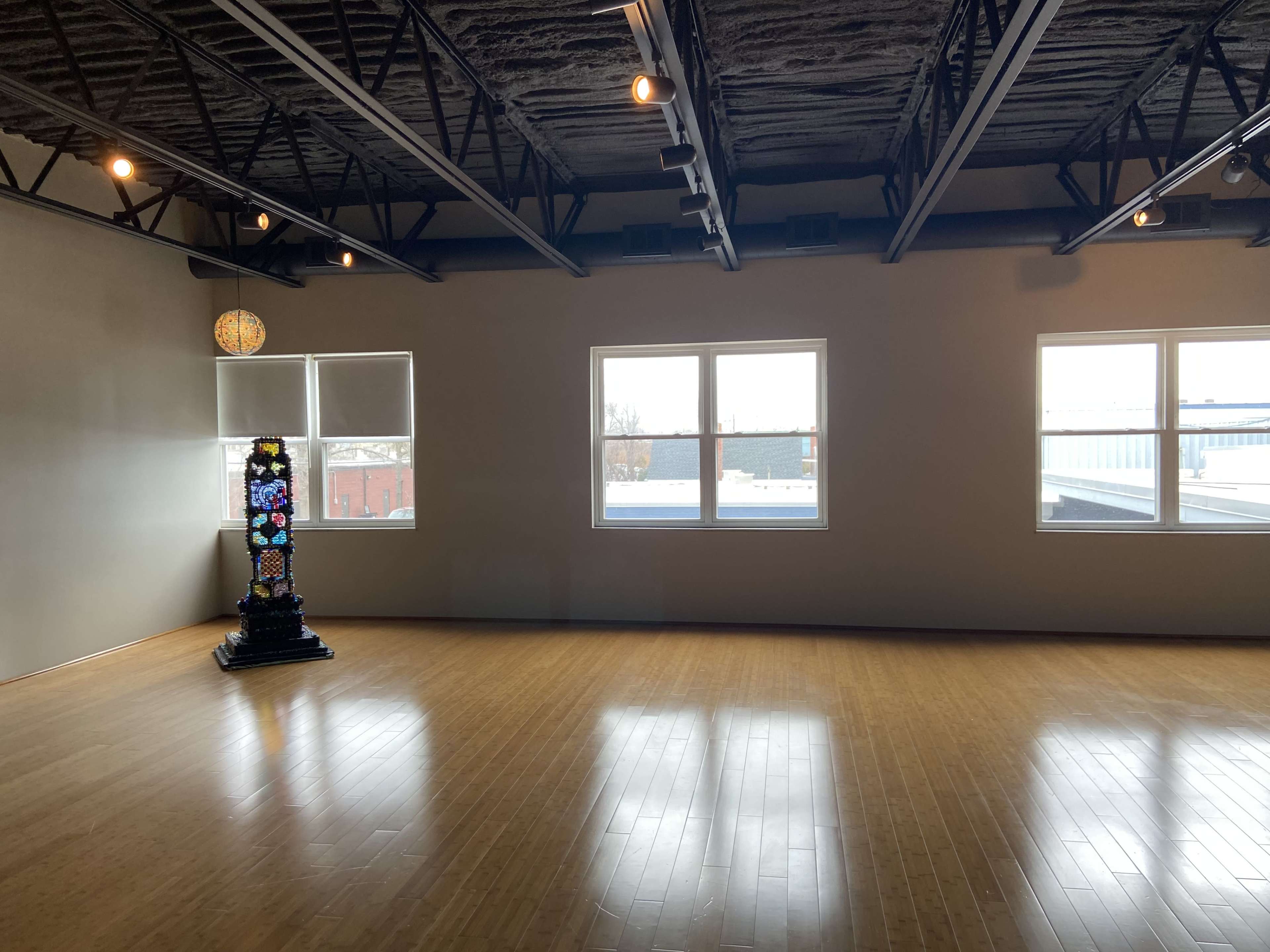 A spacious, well-lit room with large windows and polished wooden floors, featuring a decorative stand with a colorful design in one corner.