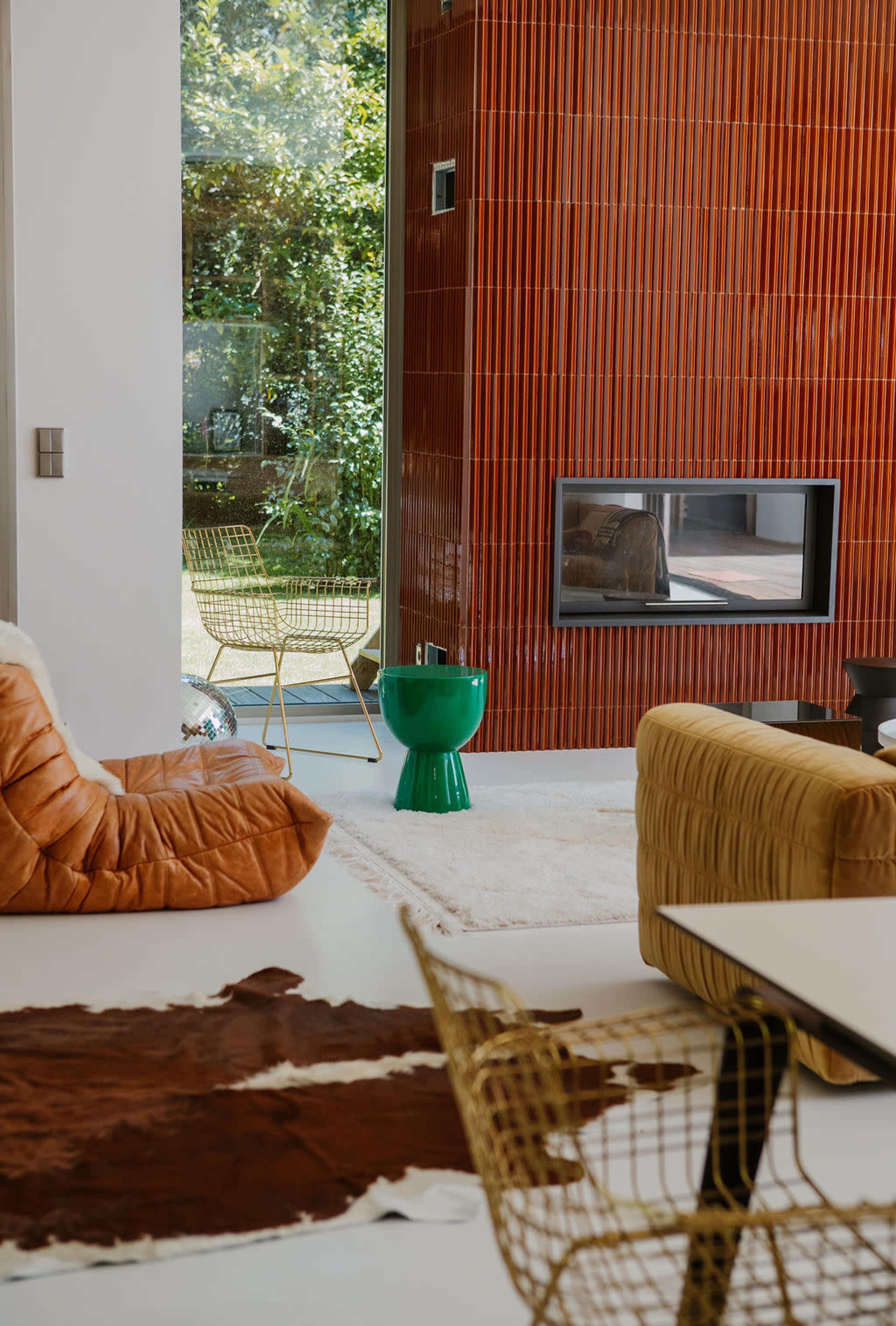 The image shows a modern living room with a fireplace, green accent table, and various seating options, including a brown lounge chair and gold wire chairs, set against a backdrop of a red textured wall.