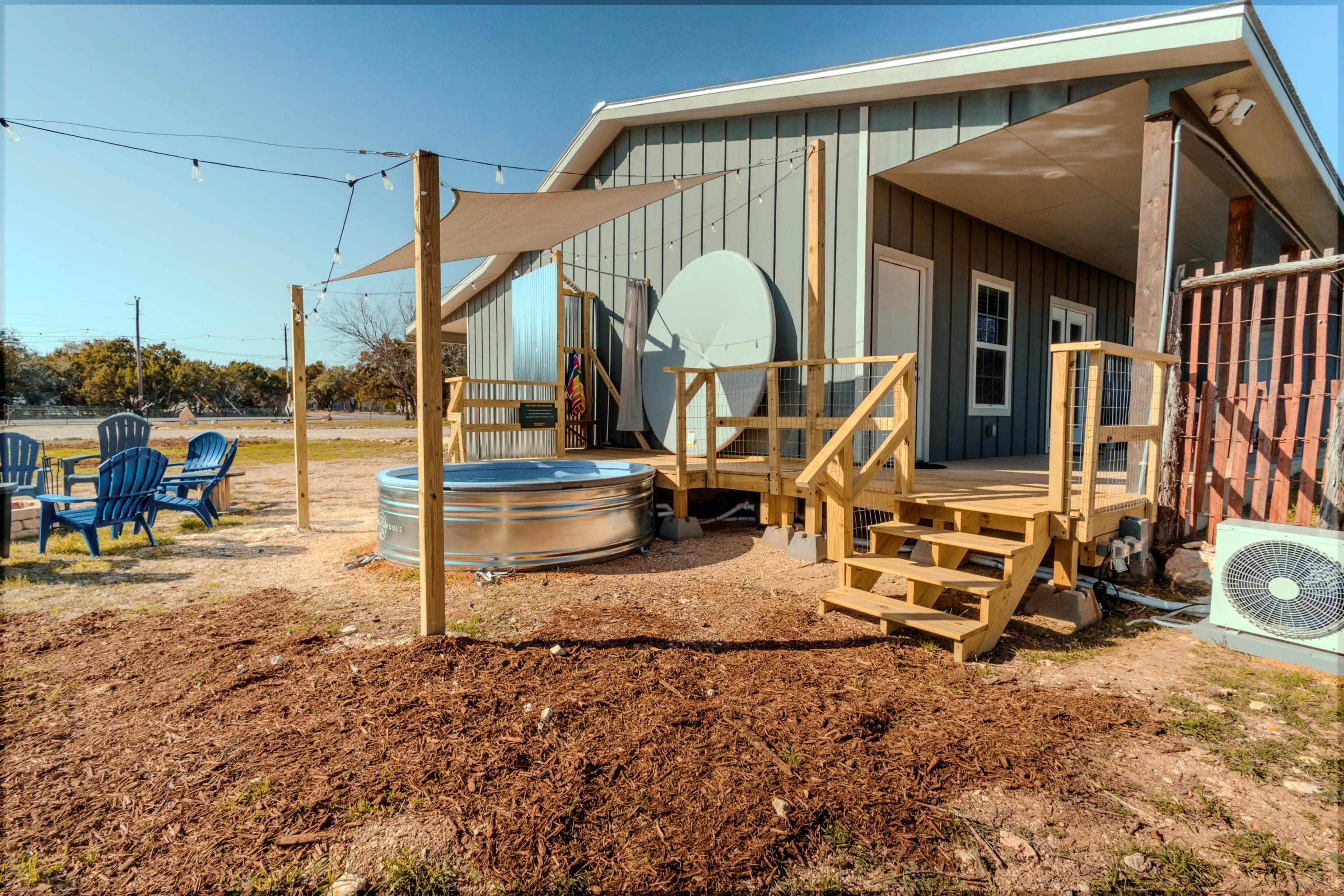 The image shows a modern outdoor area with a metal hot tub, wooden deck steps, and a shaded seating space beside a house.