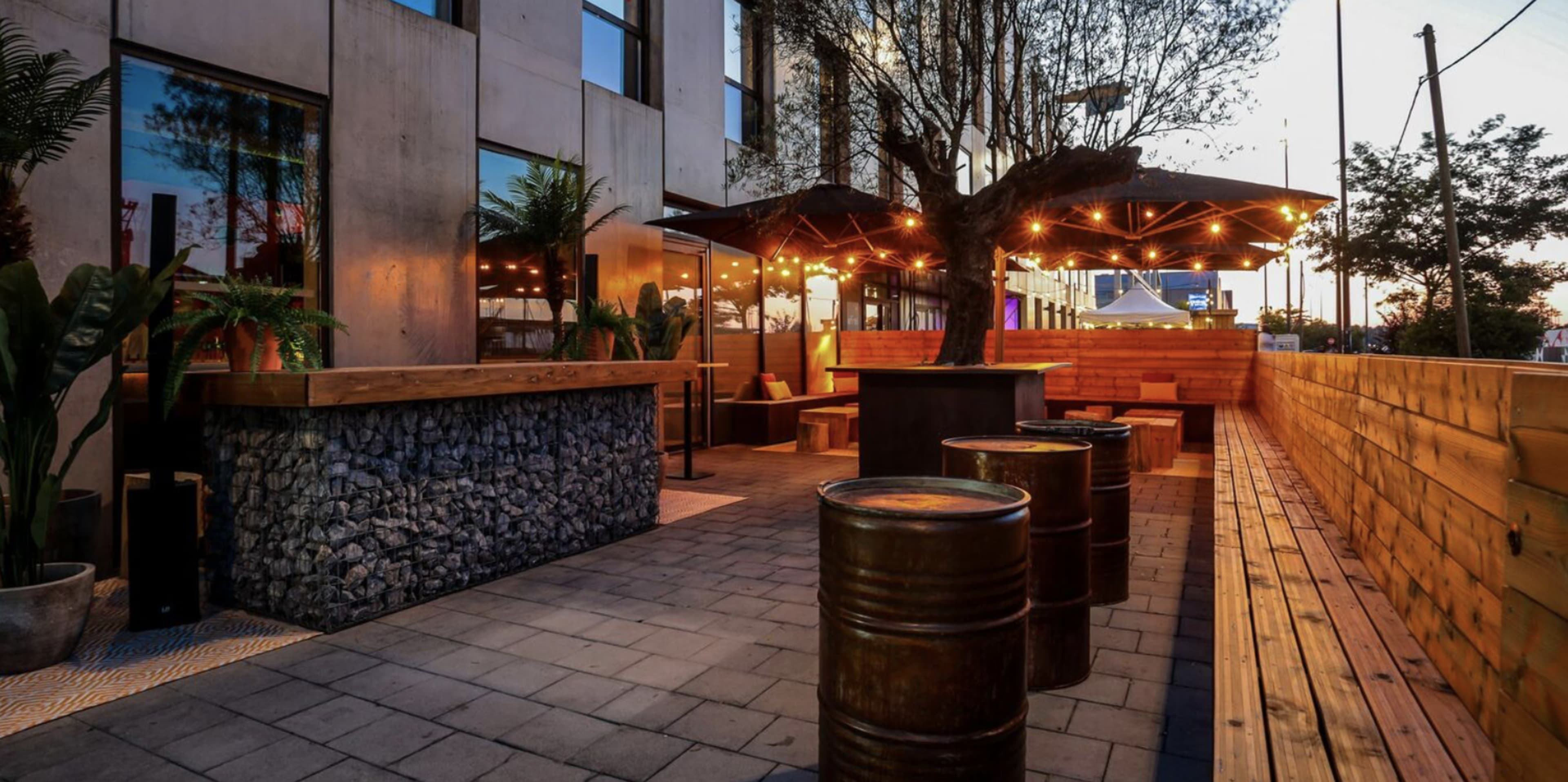 The image shows an outdoor dining area featuring wooden benches, a bar made of stone, and warm string lights illuminated against a dusk sky.