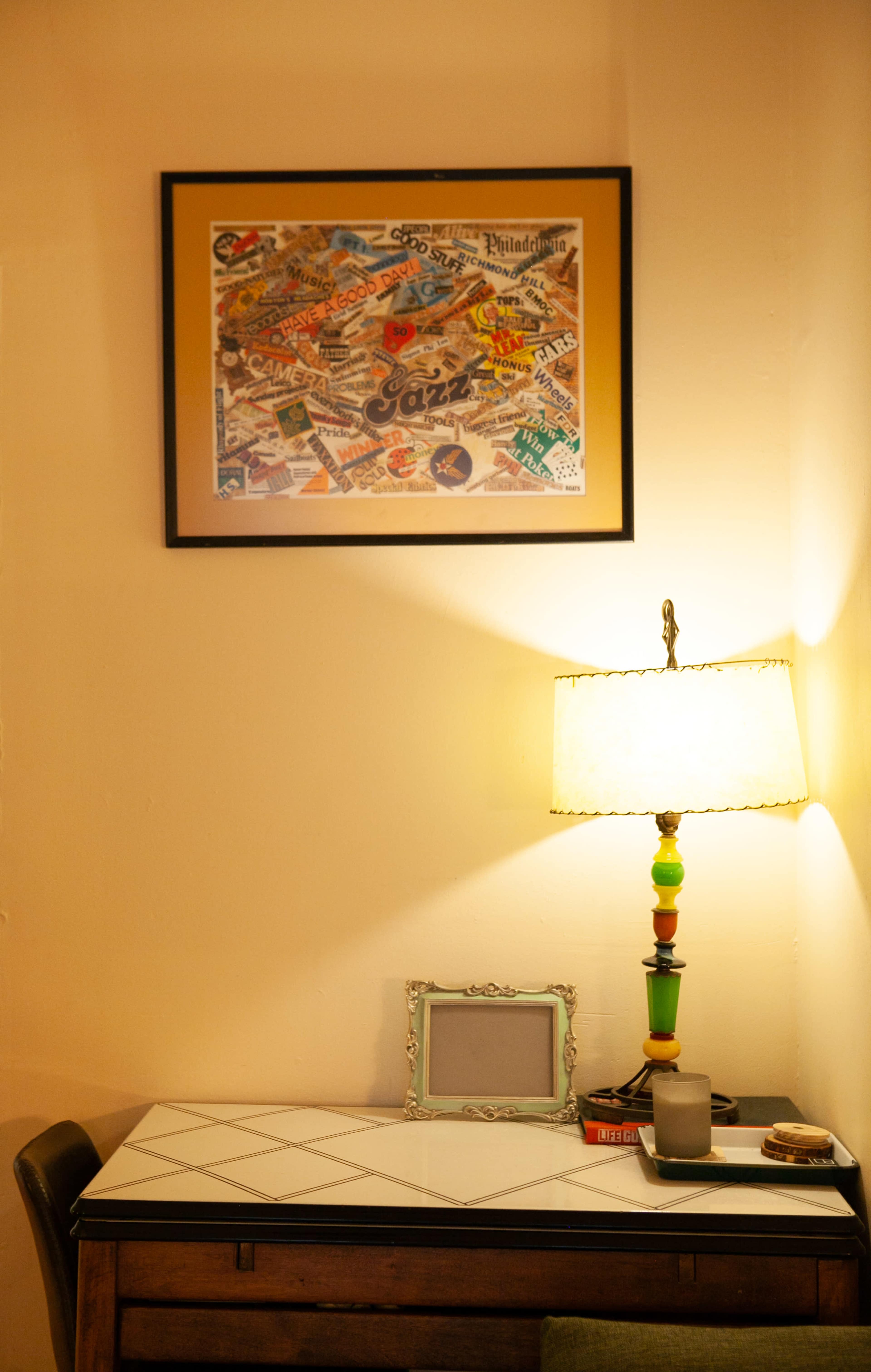 A framed collage of vintage advertisements hangs above a small desk with a lamp and decorative items.