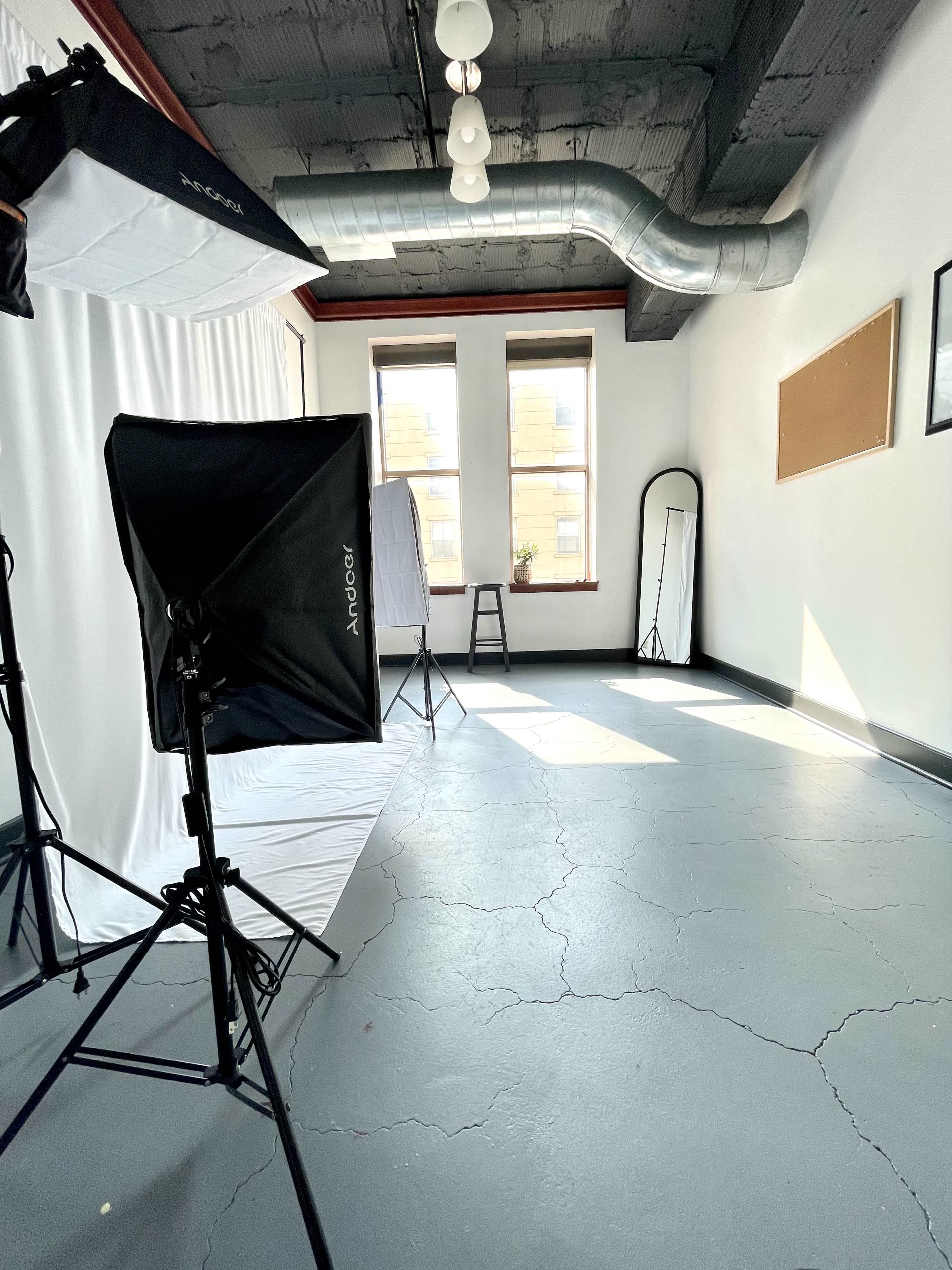 A photography studio with lighting equipment set up, a blank backdrop, and natural light streaming through windows.