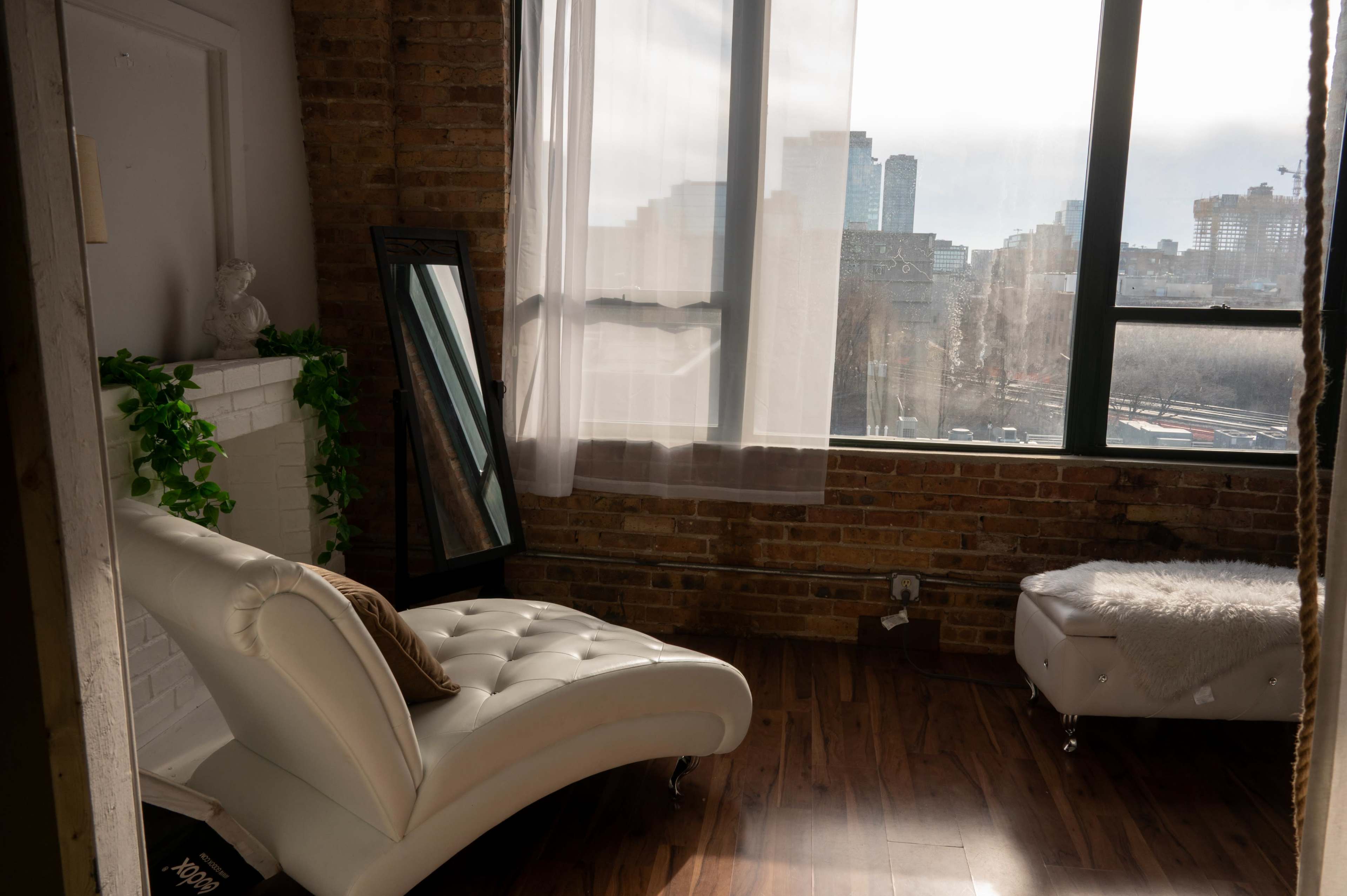 The image shows a bright room with a white chaise lounge, a large mirror, and a window revealing an urban skyline.