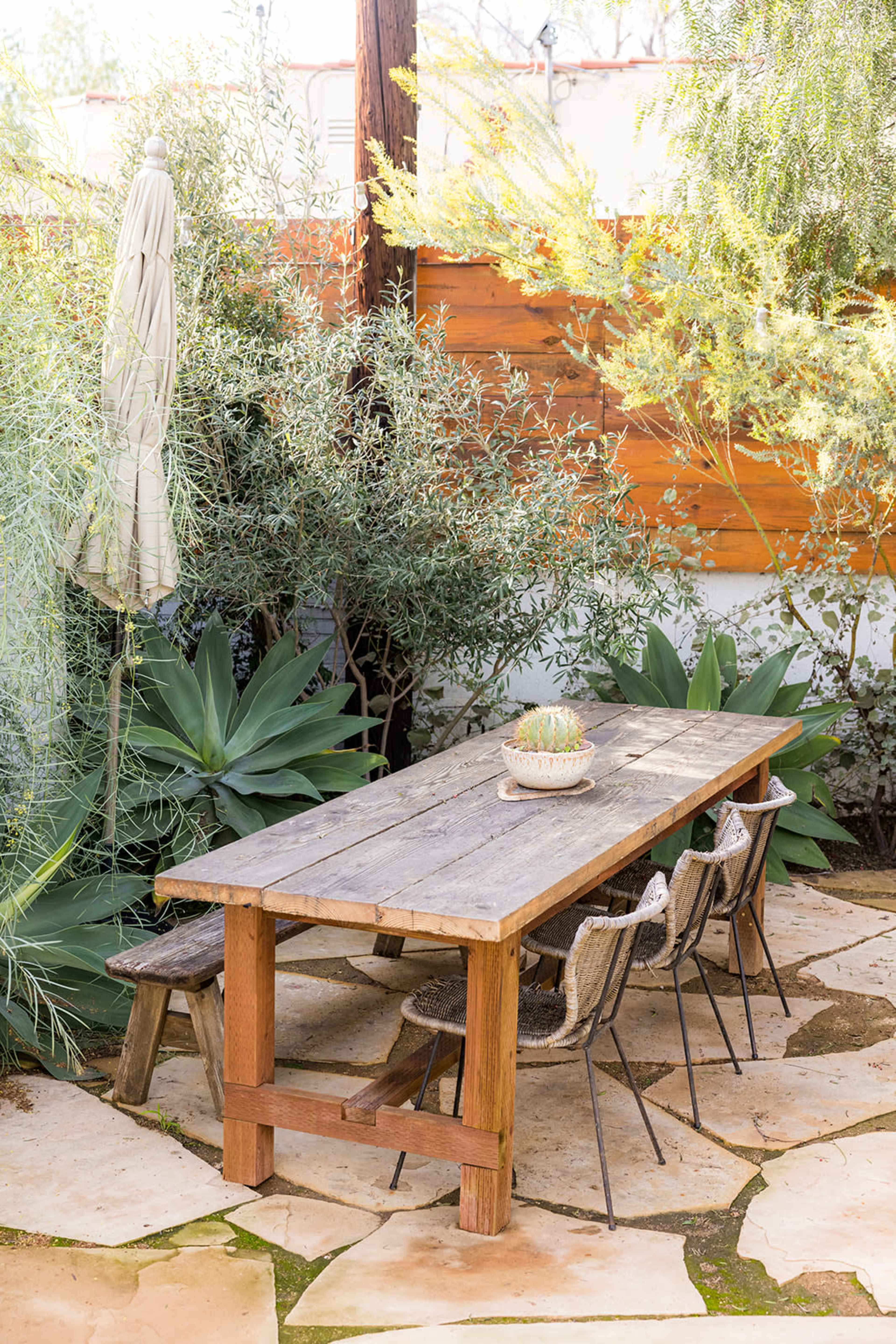 A long wooden table with chairs sits surrounded by greenery and stone pathway in a garden setting.