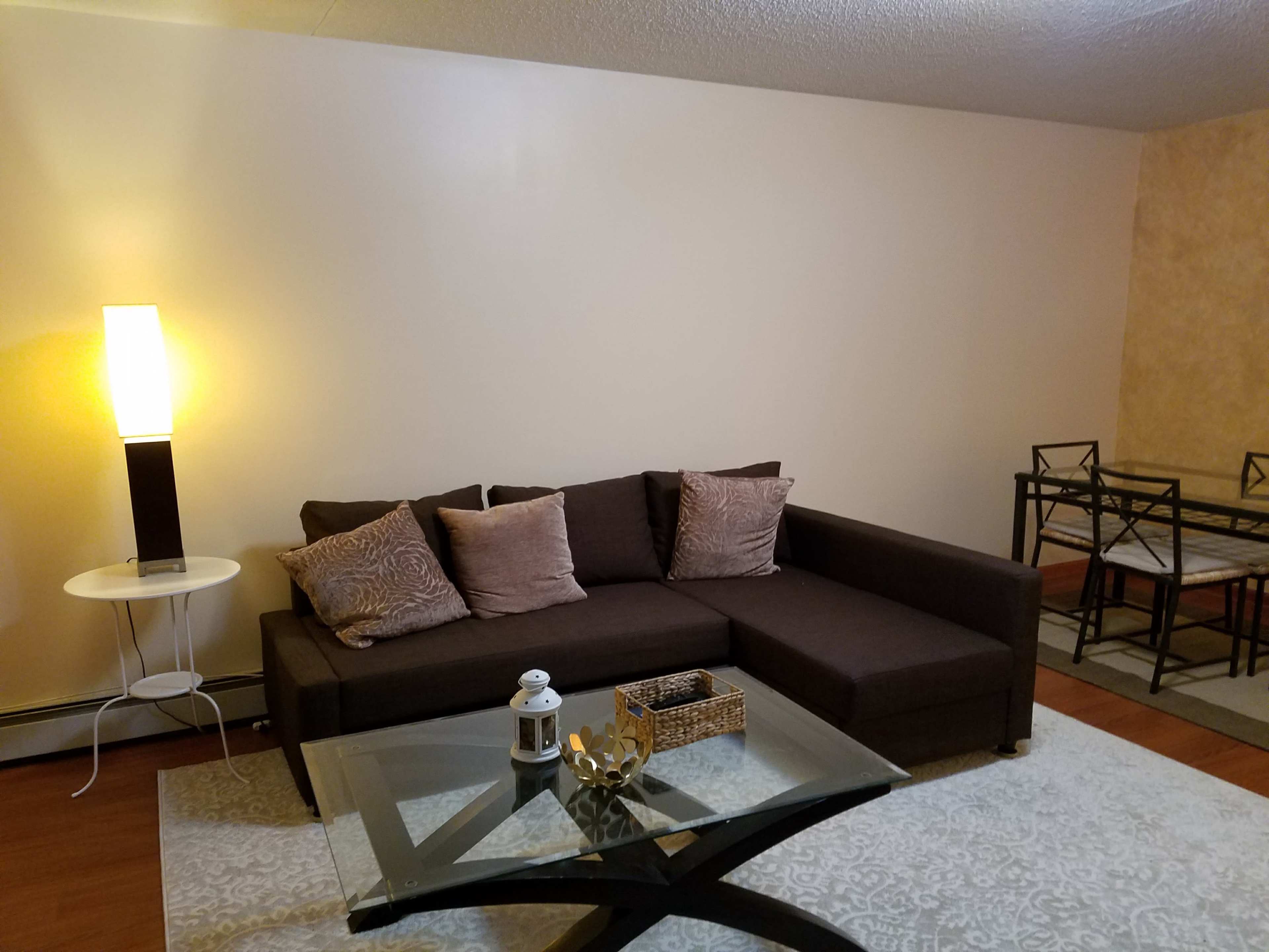 The image shows a modern living room featuring a dark sectional sofa with pillows, a glass coffee table, and a small round side table with a lamp.