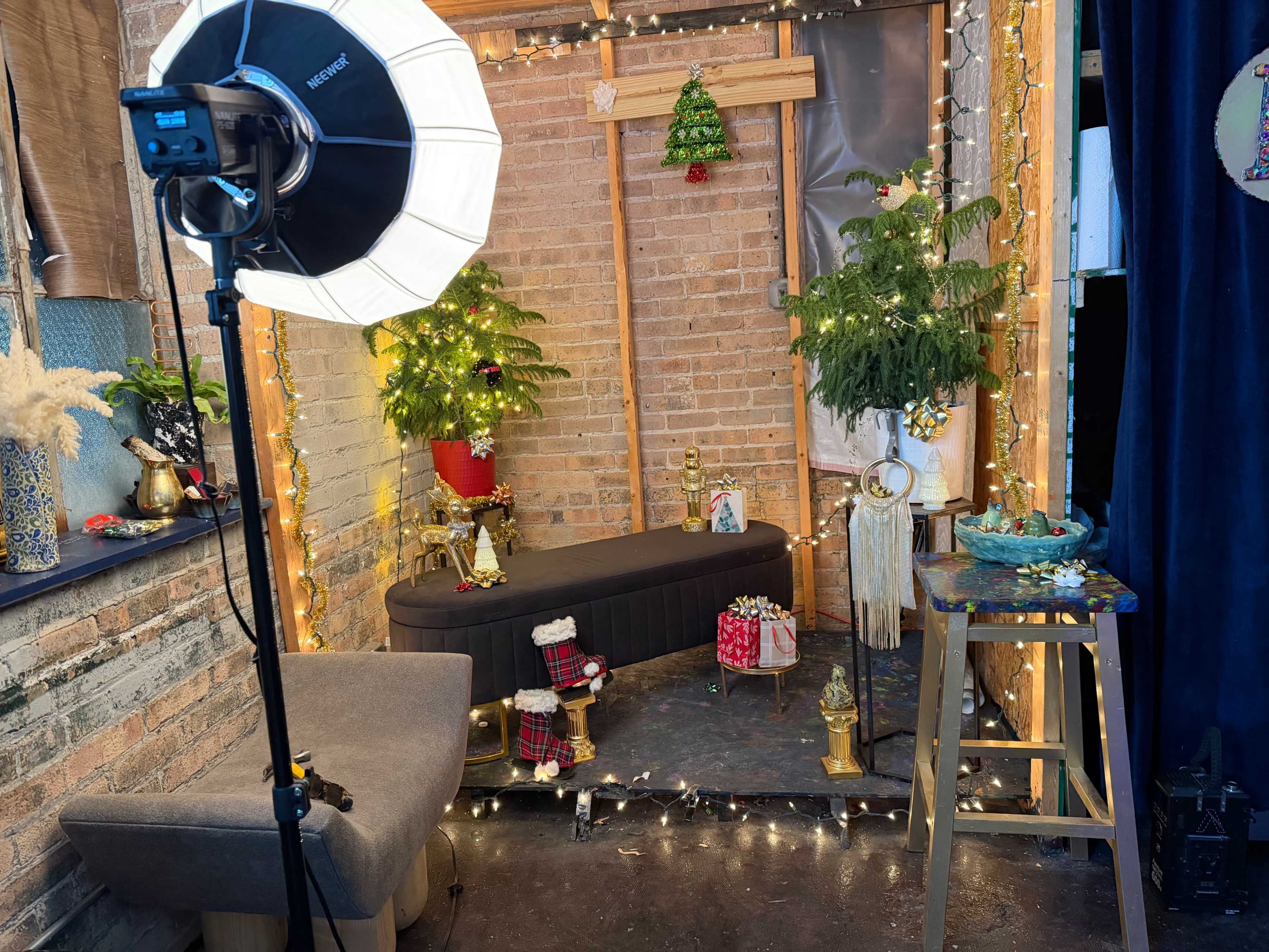 The image shows a festive photo studio setup with a decorated corner featuring a couch, small Christmas trees, lights, and holiday-themed decorations.