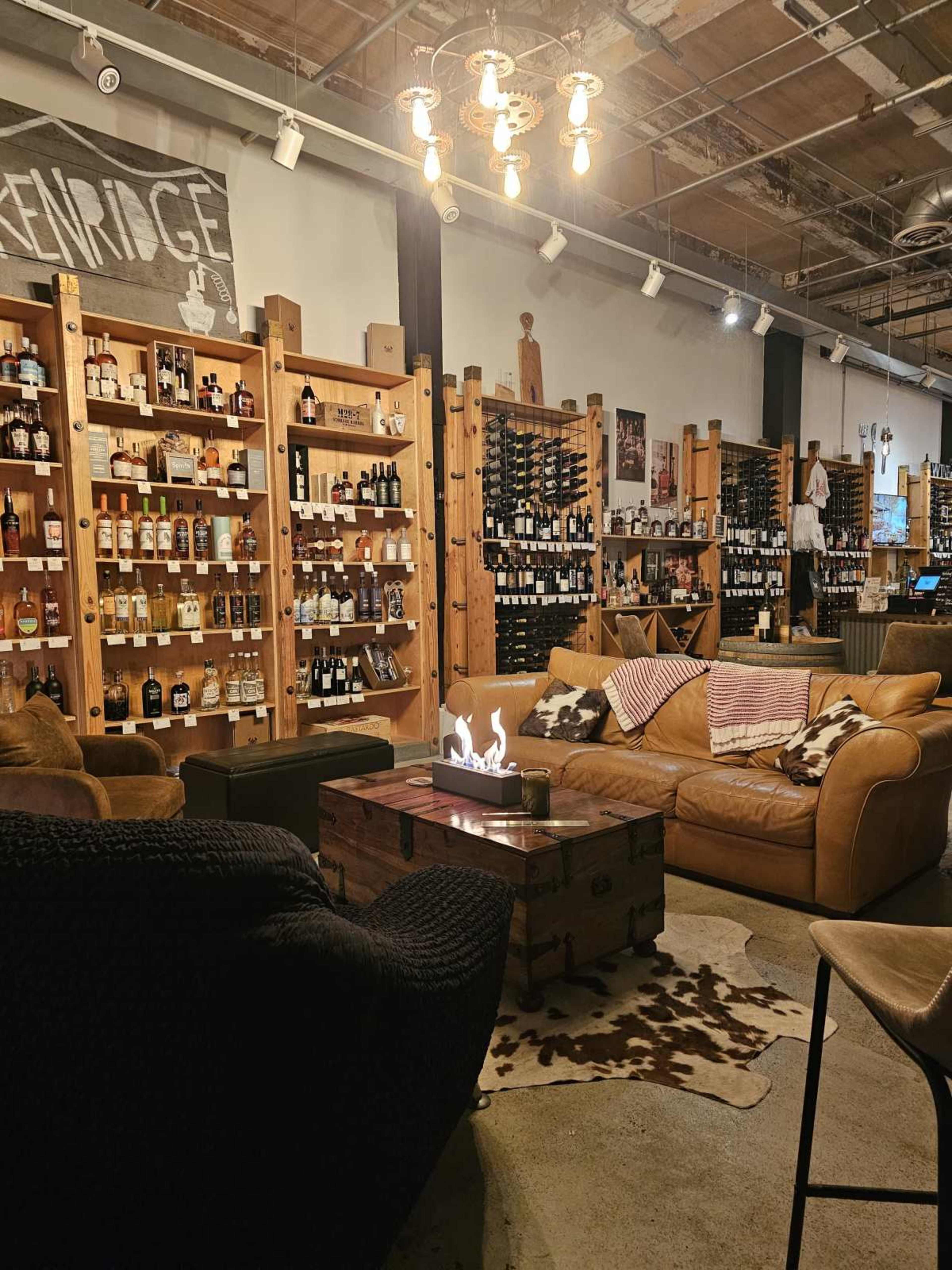 The image shows a cozy liquor store interior with wooden shelves filled with various bottles and comfortable seating arrangements featuring leather sofas and a decorative coffee table.