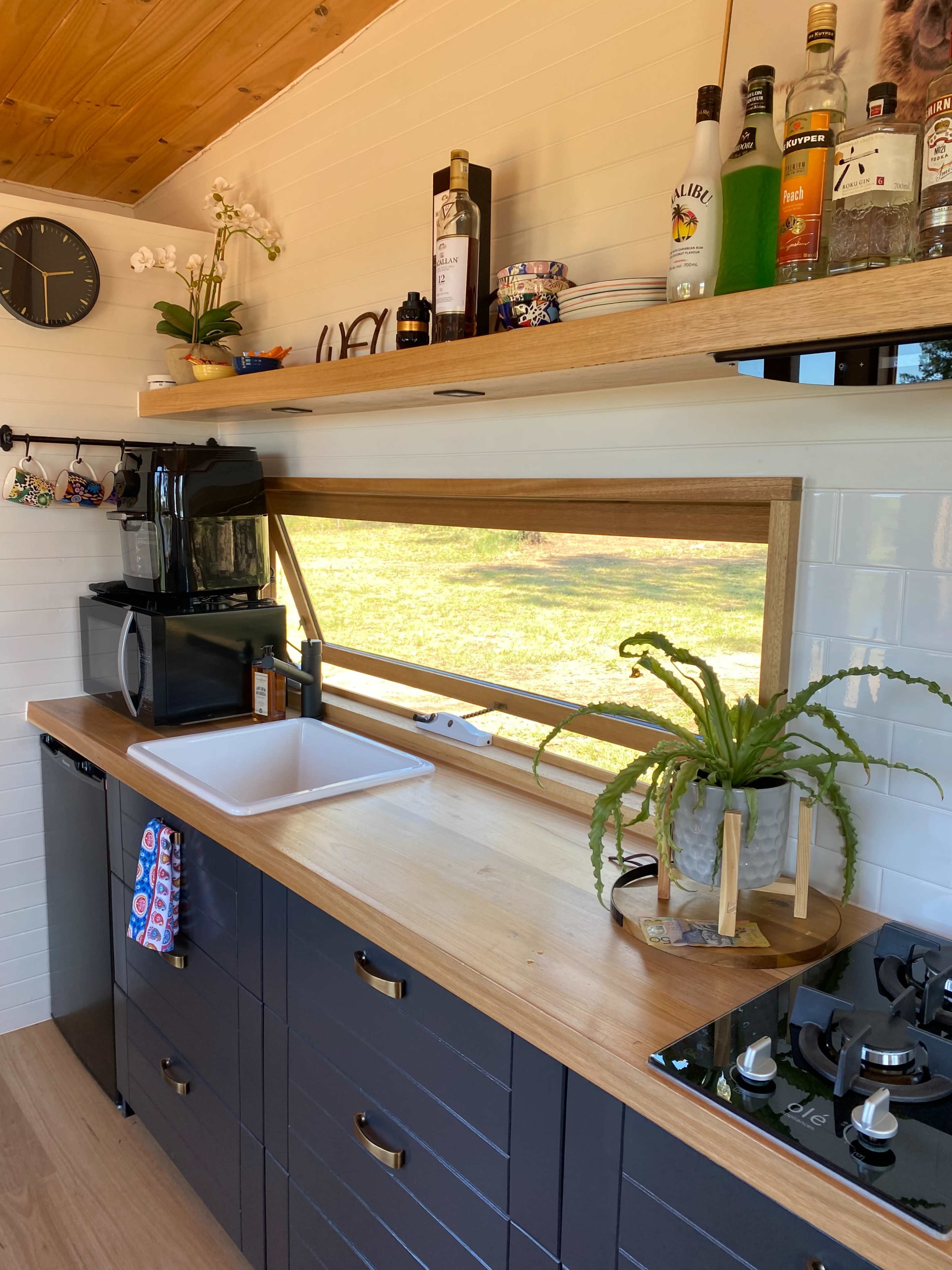 A kitchen with a wooden countertop, a sink, a coffee maker, and a window overlooking an outdoor view, along with shelving displaying drinks and decorative items.