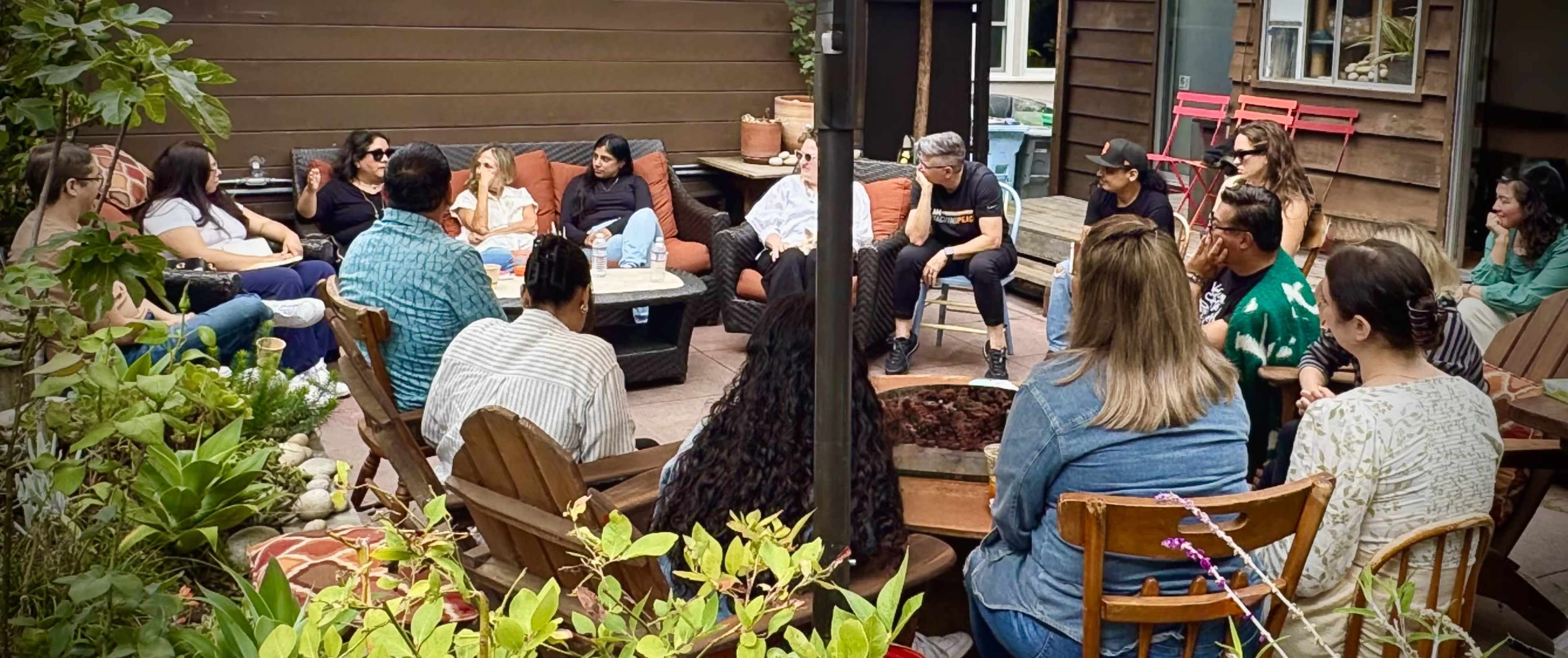 A group of people sits in an outdoor gathering area, engaged in conversation around a central fire pit.