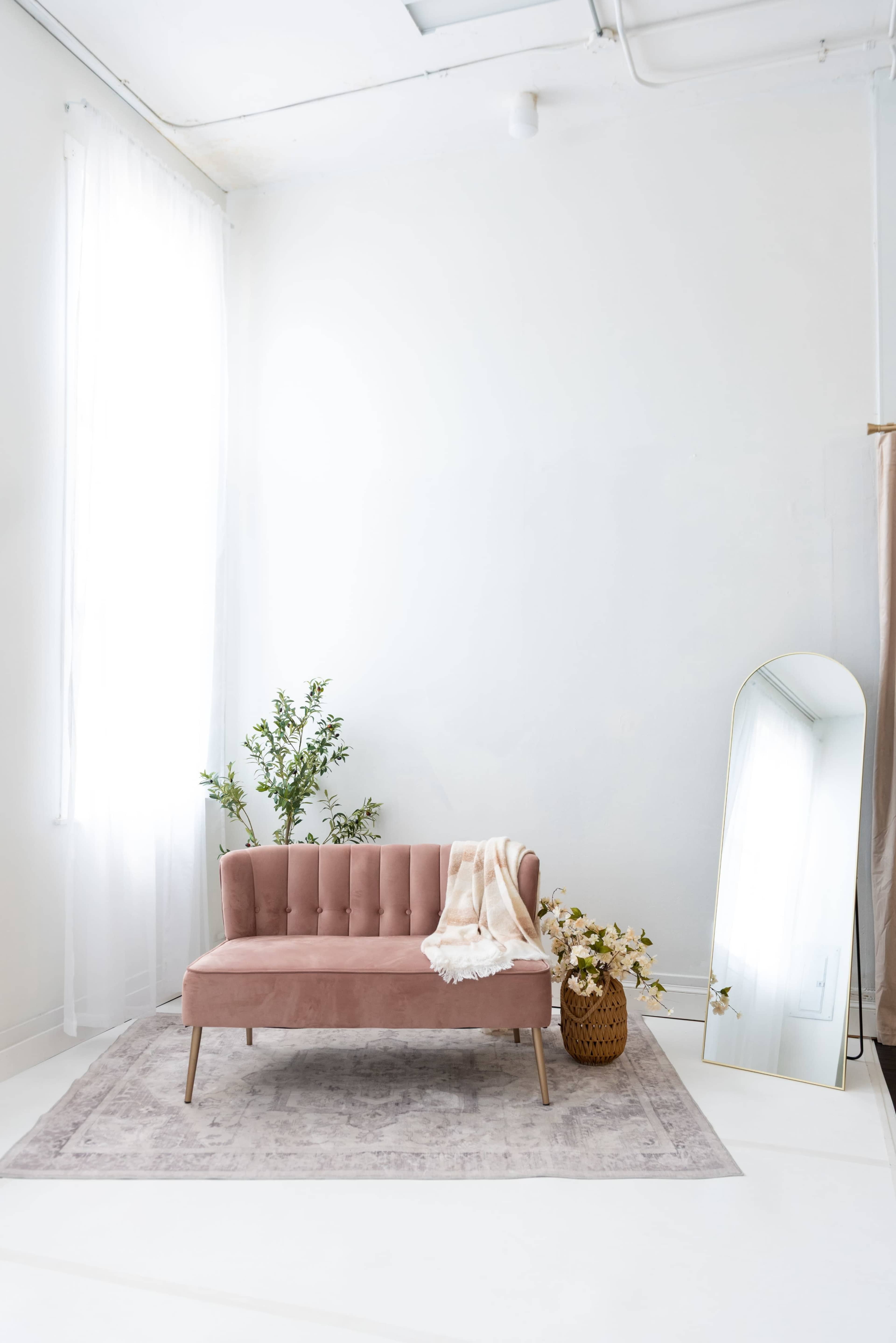 A pink velvet sofa is positioned in a bright, minimalist room with a large mirror and potted plants.