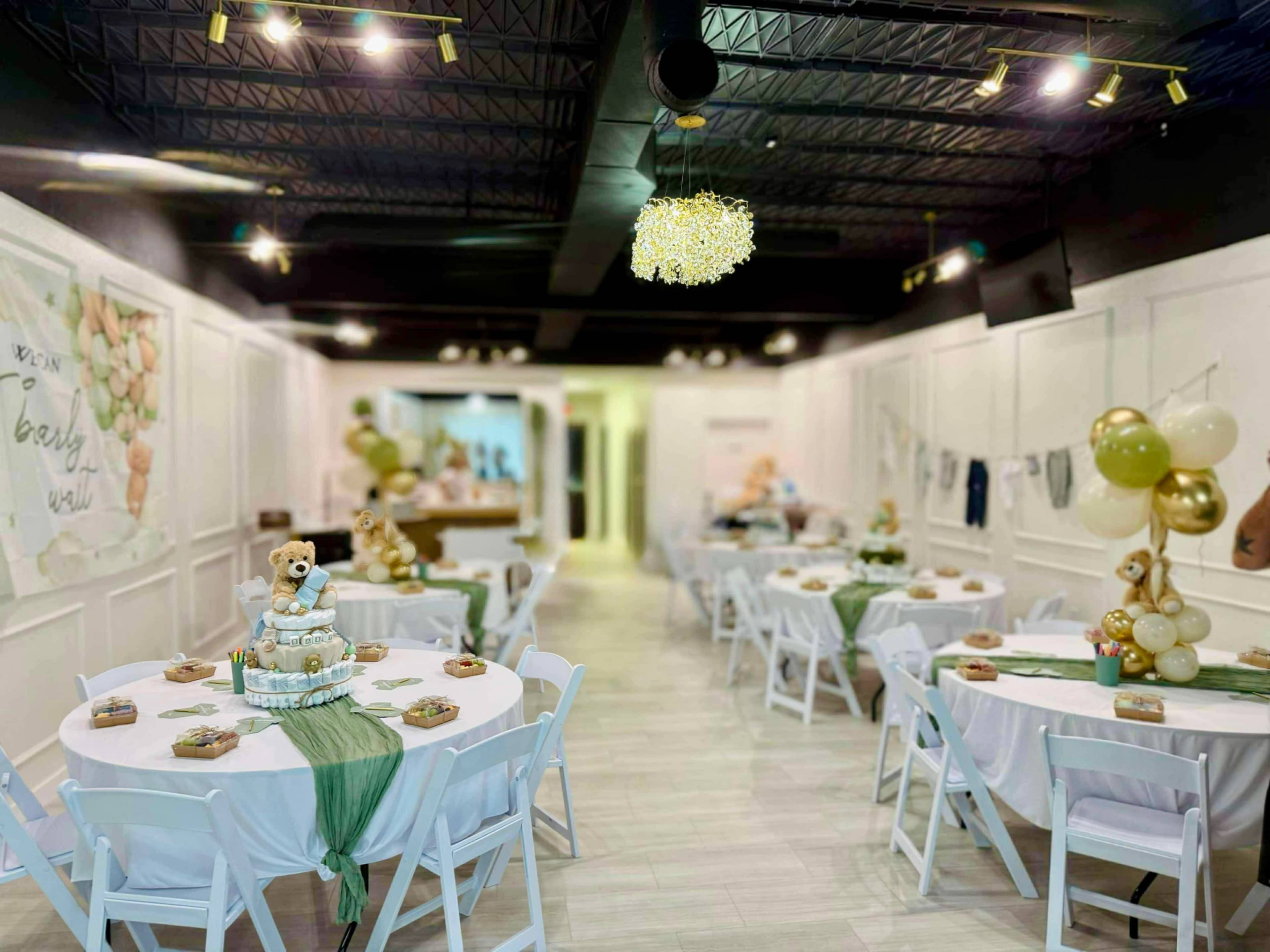 The image depicts a decorated event space with white tables set for a celebration, featuring a cake and balloons, amidst a light and airy ambiance.
