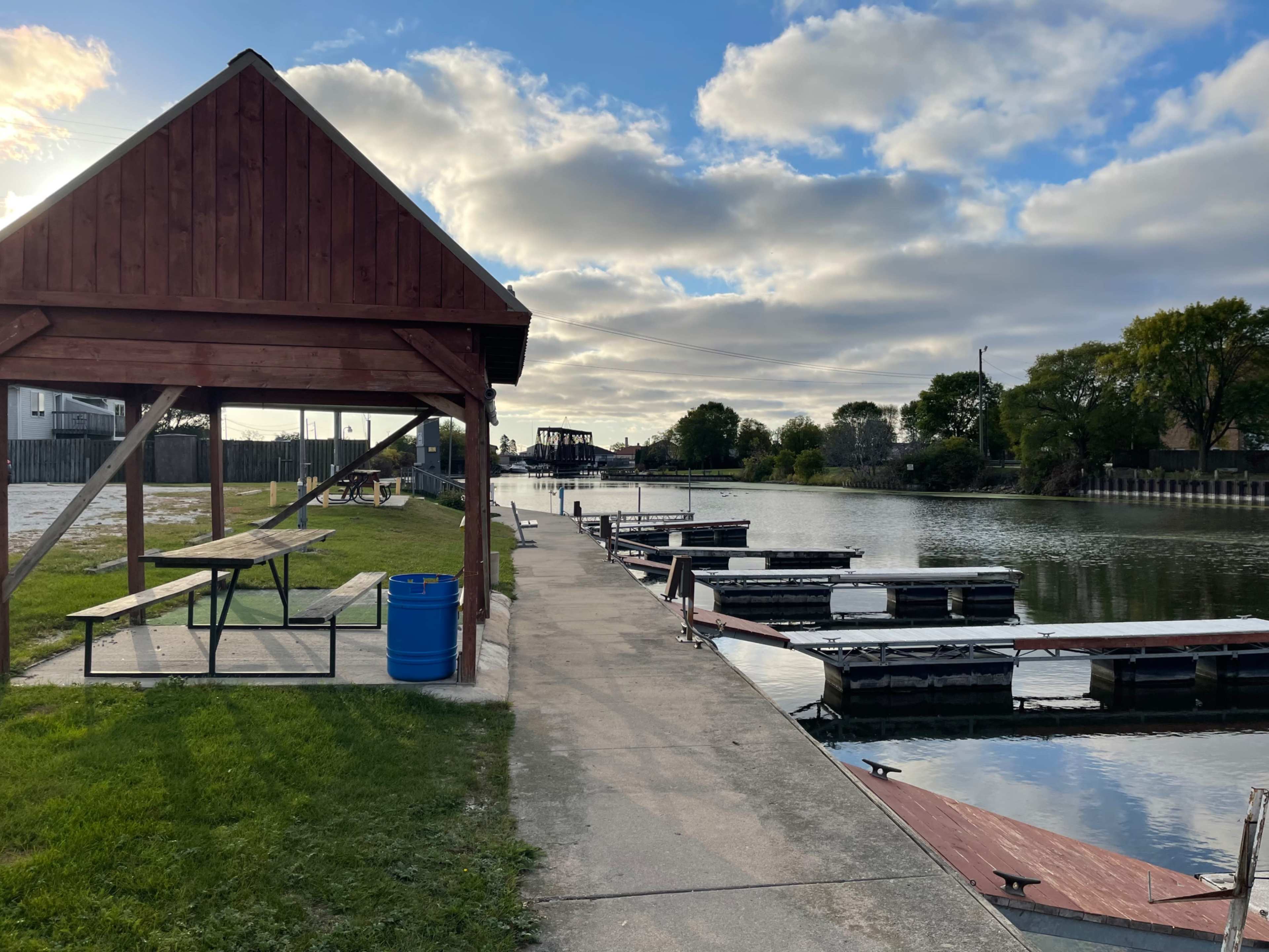 A wooden gazebo alongside a walkway extending to a marina with several docked boats on a calm waterway.