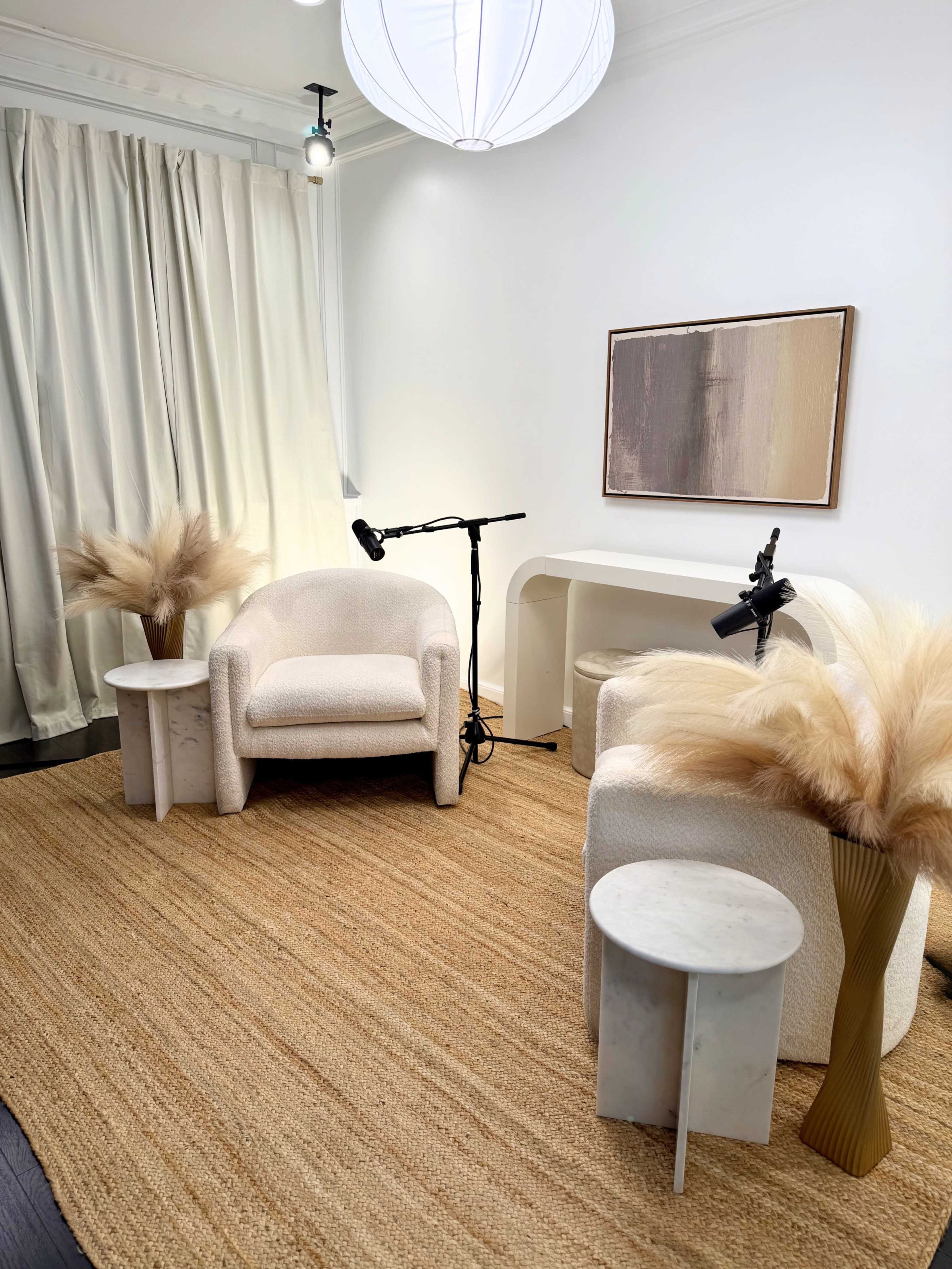 The image features a cozy interior with two modern armchairs, a small round table, a console table, and decorative arrangements of dried plants, all set against light-colored walls and soft curtains.