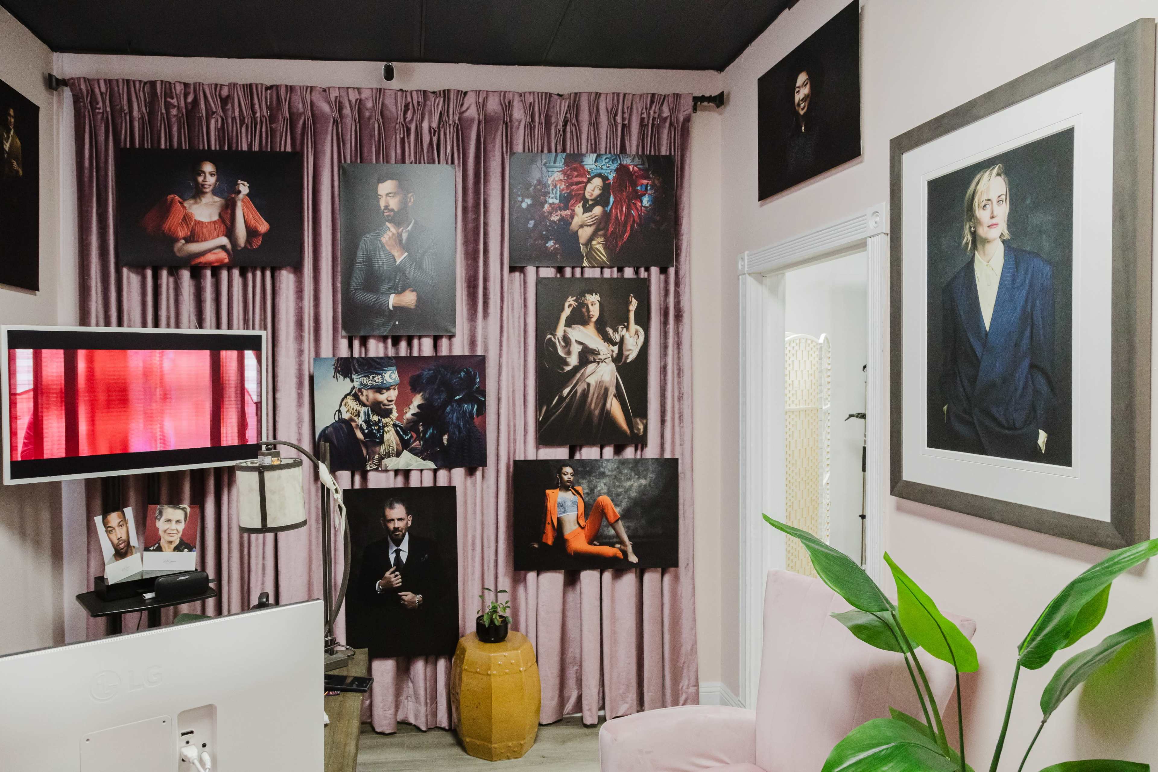 The image shows a styled room with pink walls, featuring various framed portraits on the curtains and walls, alongside a television and a decorative plant.