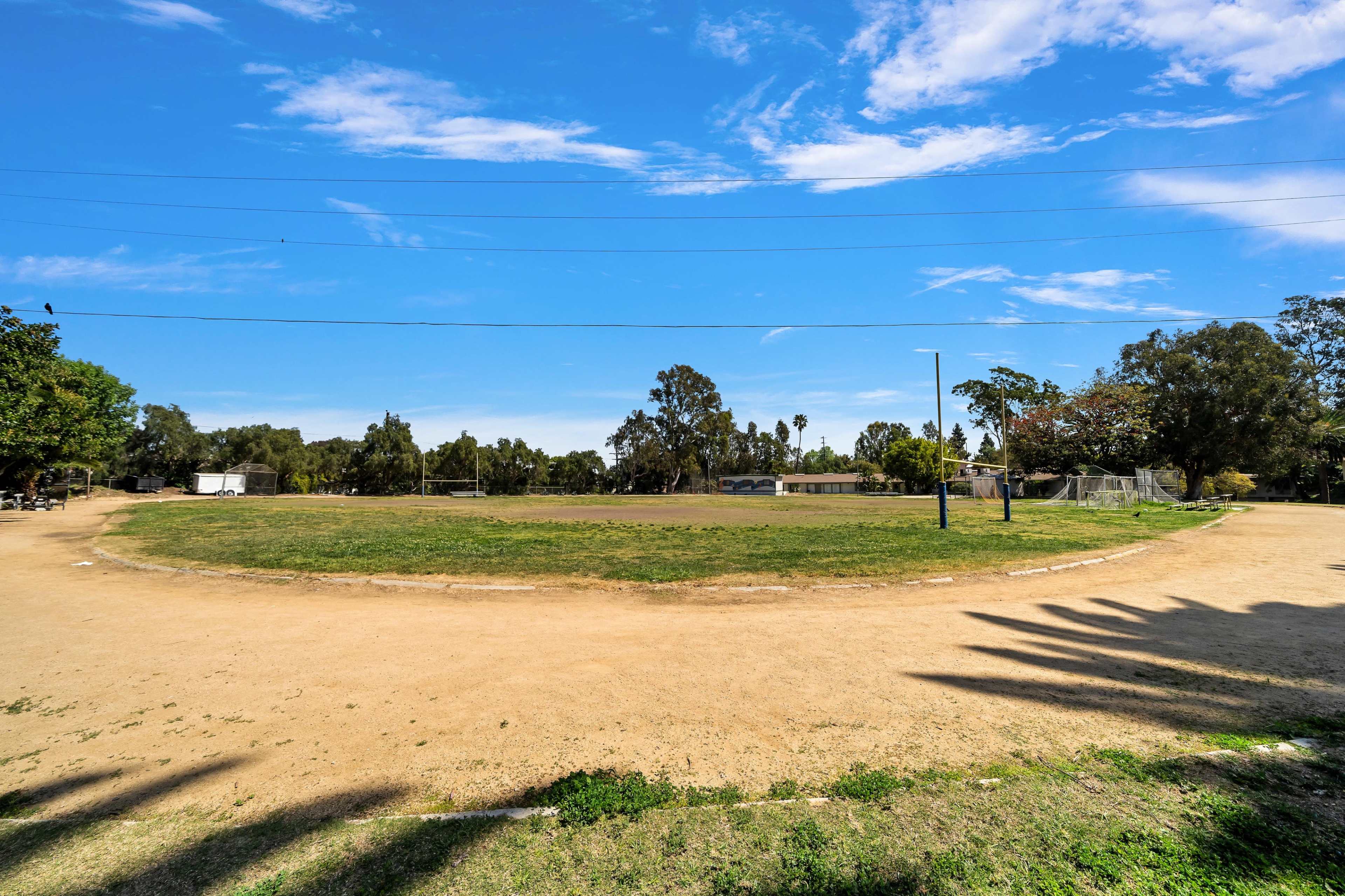A dirt track curves around a grassy area in a park, with trees and a soccer field visible in the background.