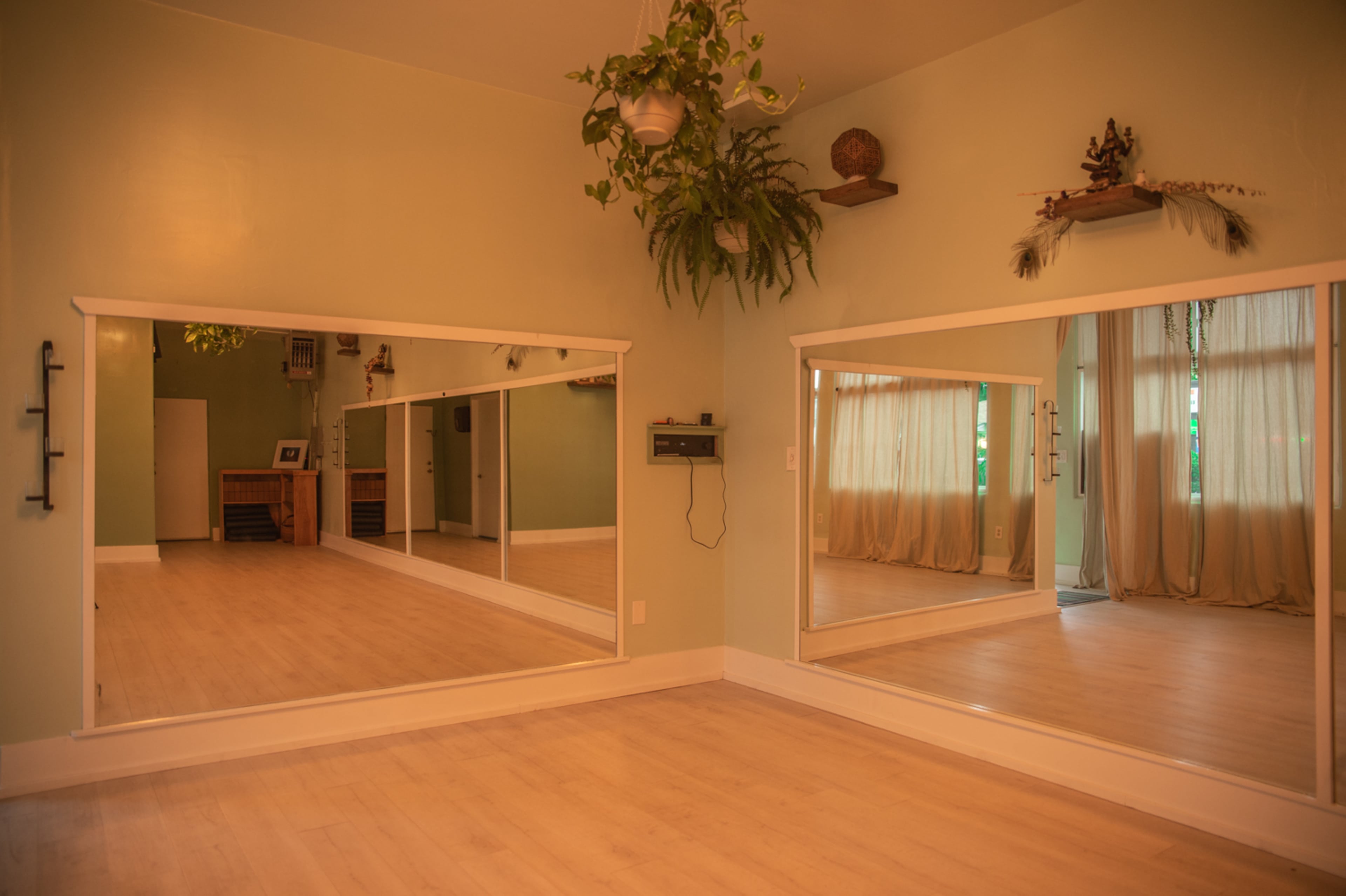 The image shows a spacious room with light green walls, large mirrors along one side, wooden flooring, and natural light filtering through sheer curtains.
