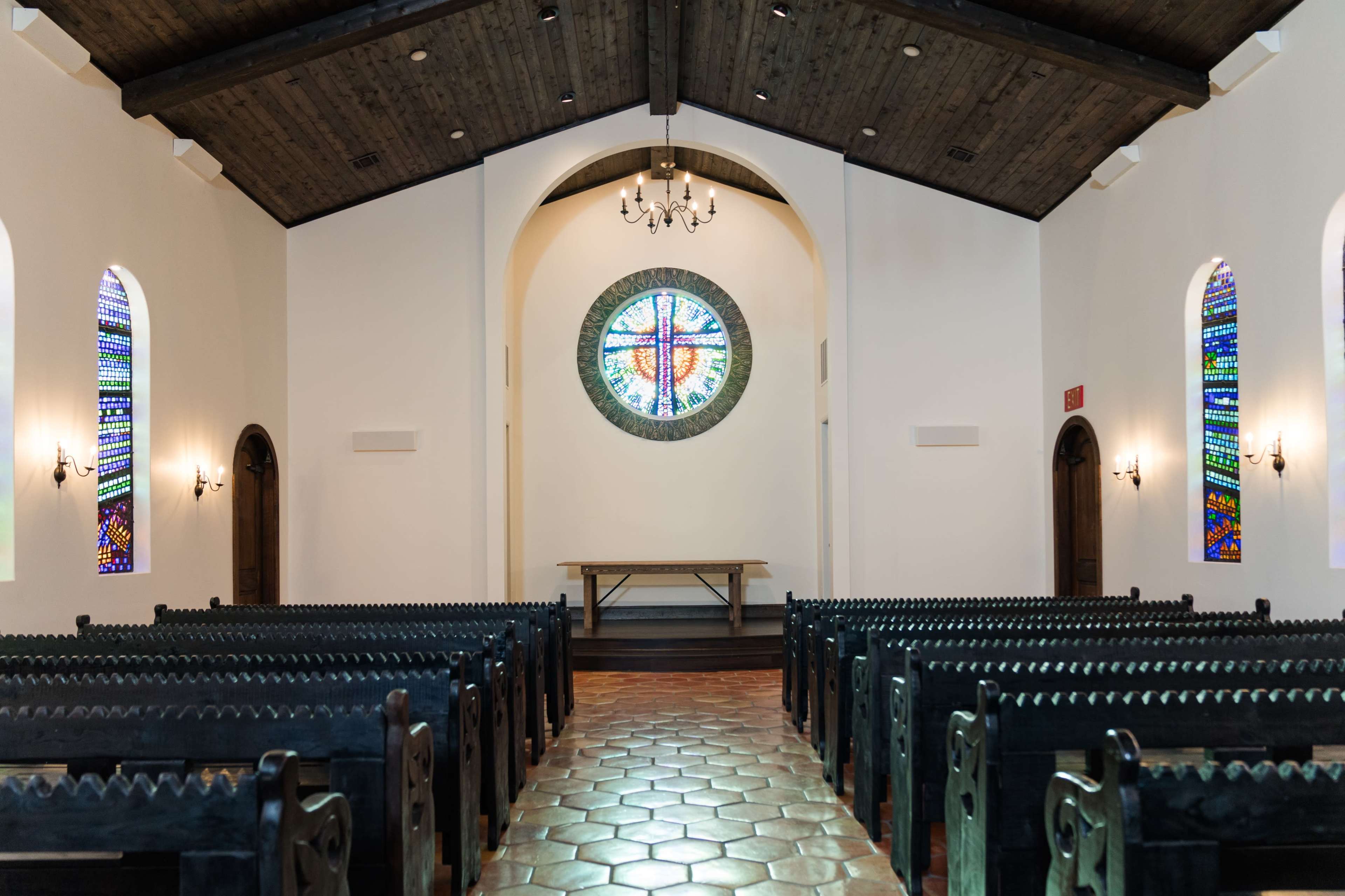 The interior of a church featuring rows of wooden pews, a central circular stained glass window, and a simple altar table at the front.