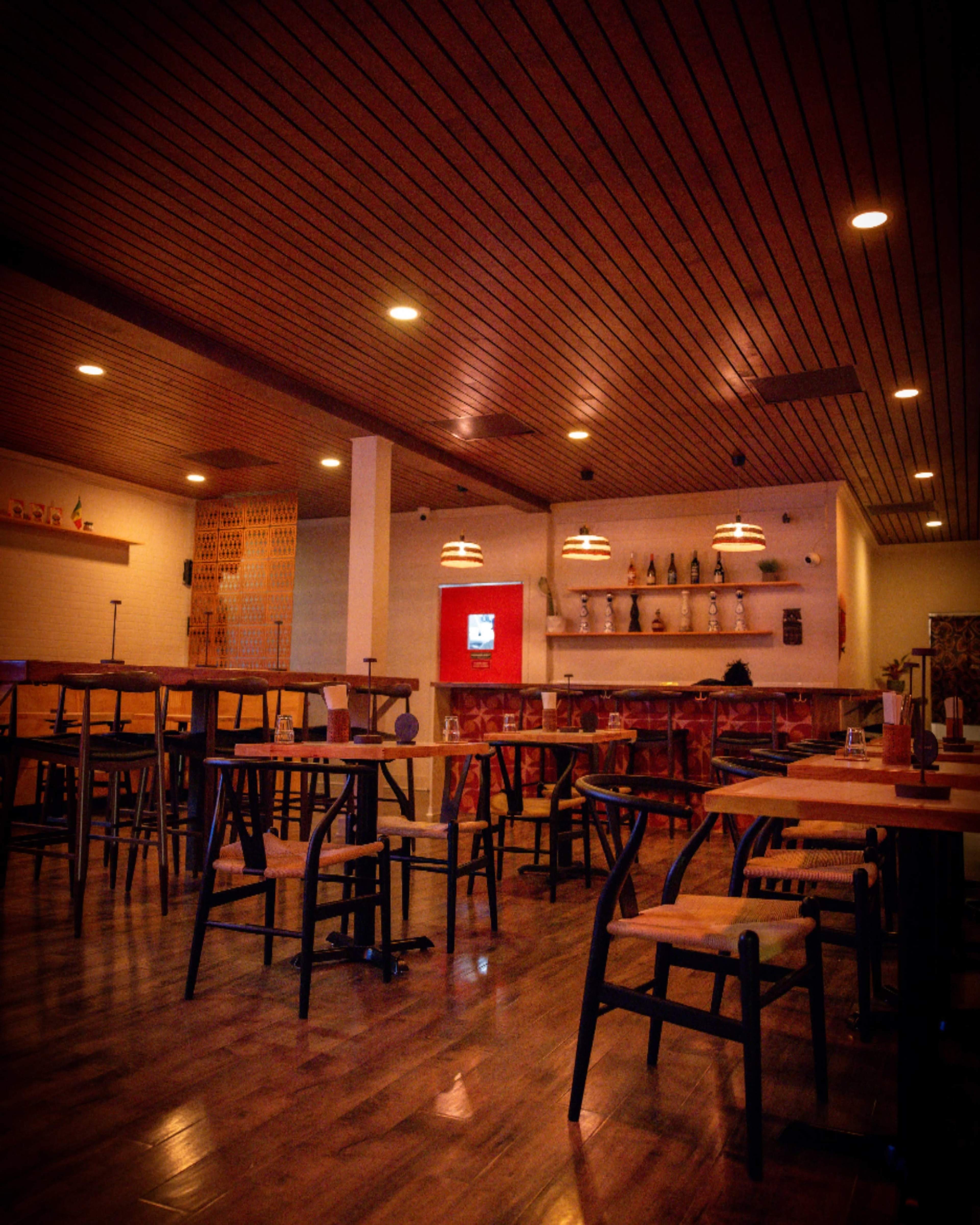 The image shows an empty restaurant with wooden tables and chairs, warm lighting, and a minimalist decor style.