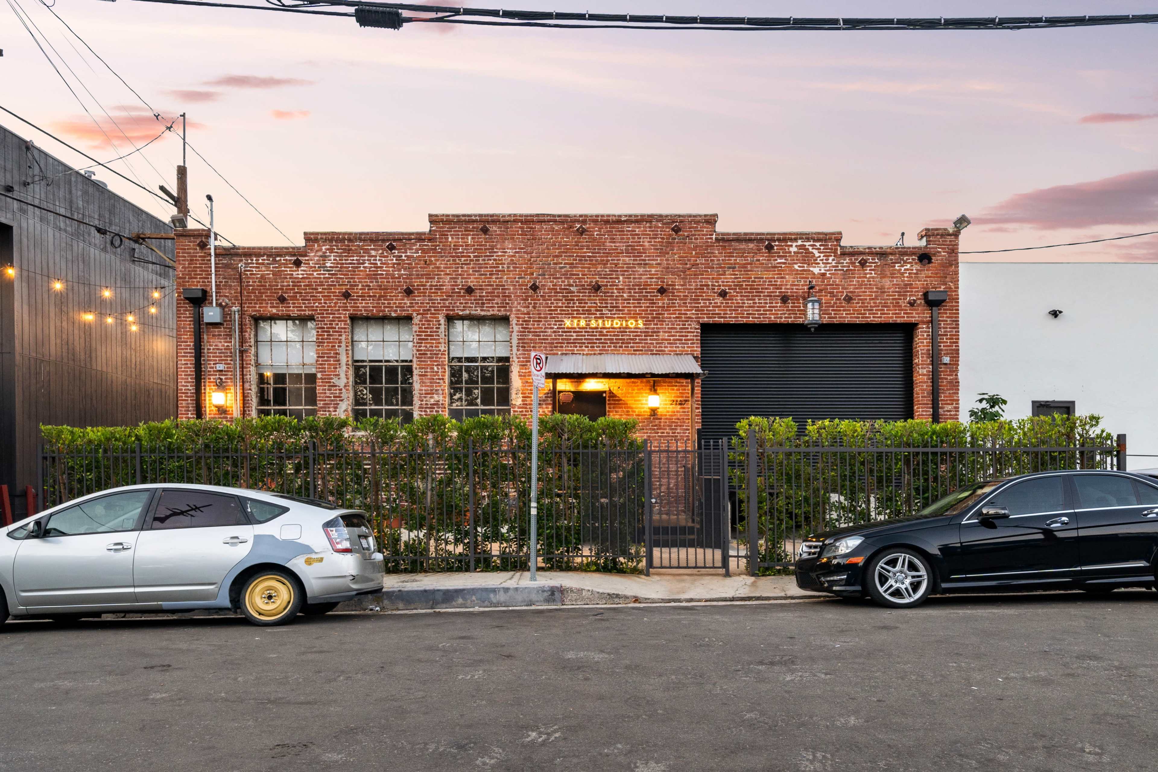 A brick building with large windows and a black roll-up door, surrounded by a low green hedge and parked cars on a street.
