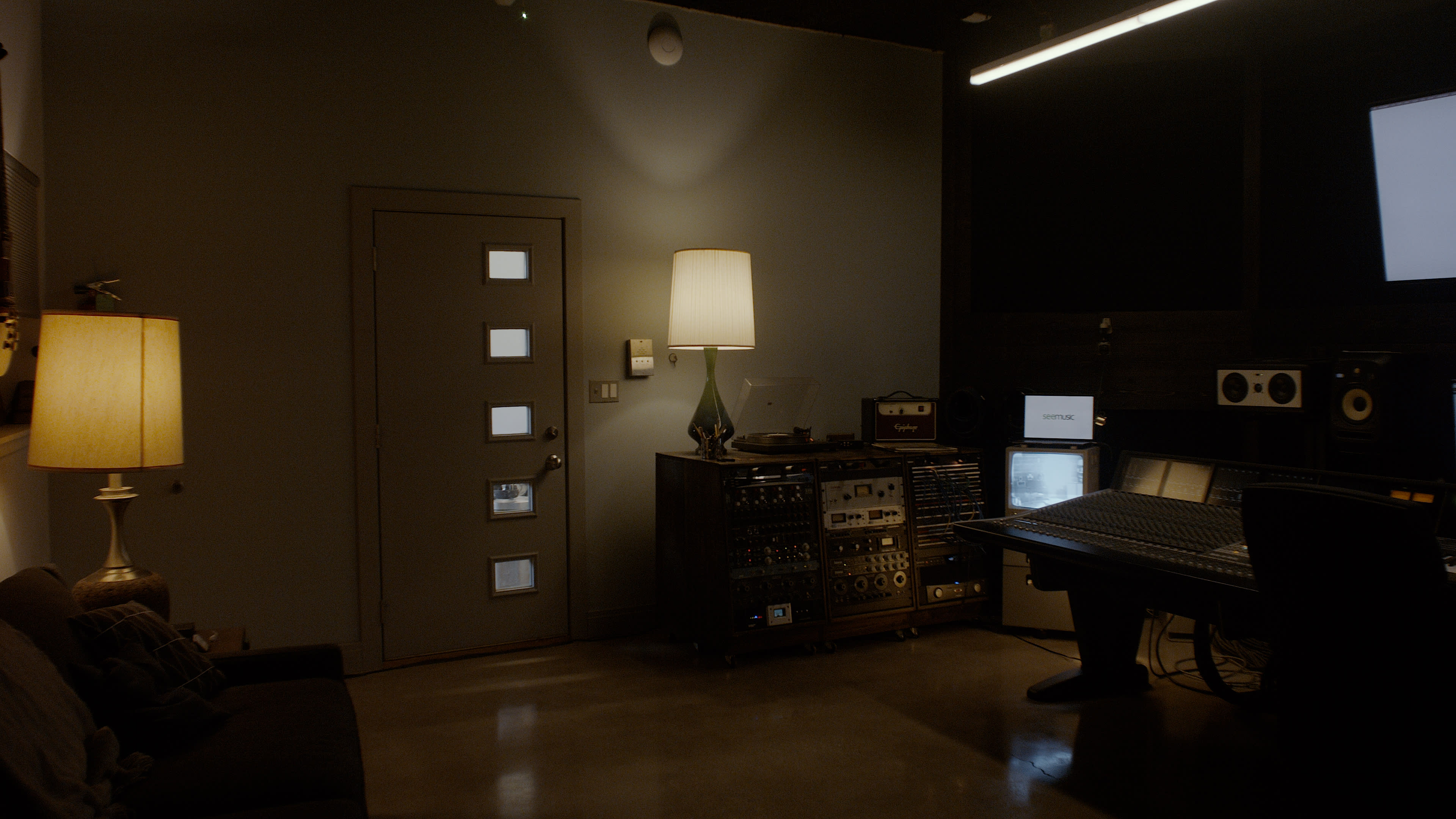 The image shows a dimly lit recording studio featuring a sofa, a lamp, a console with audio equipment, and a door with multiple windows.