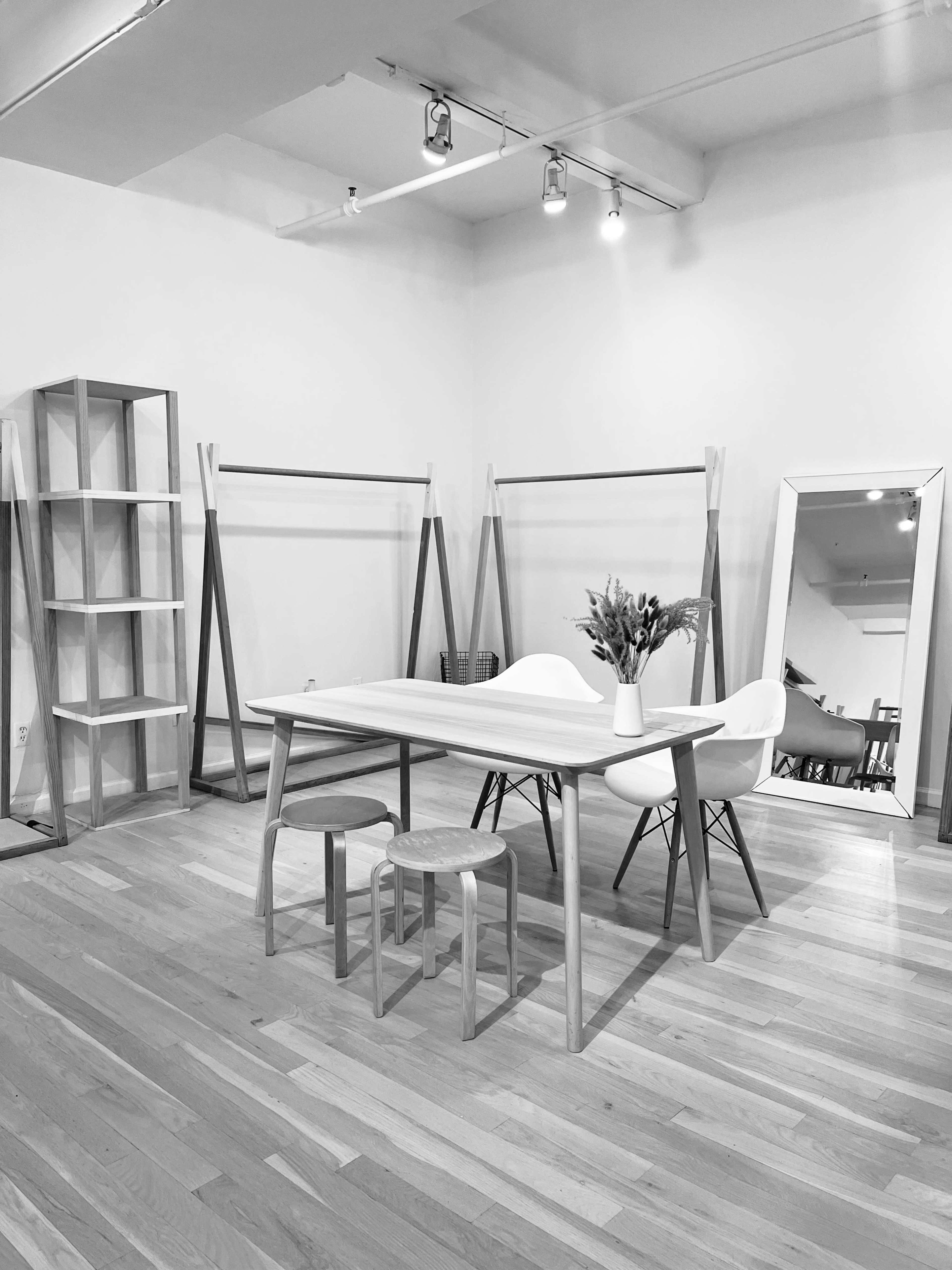 A minimalist room with a wooden table surrounded by three stools, a tall shelving unit, clothes racks, and a mirror against the wall.