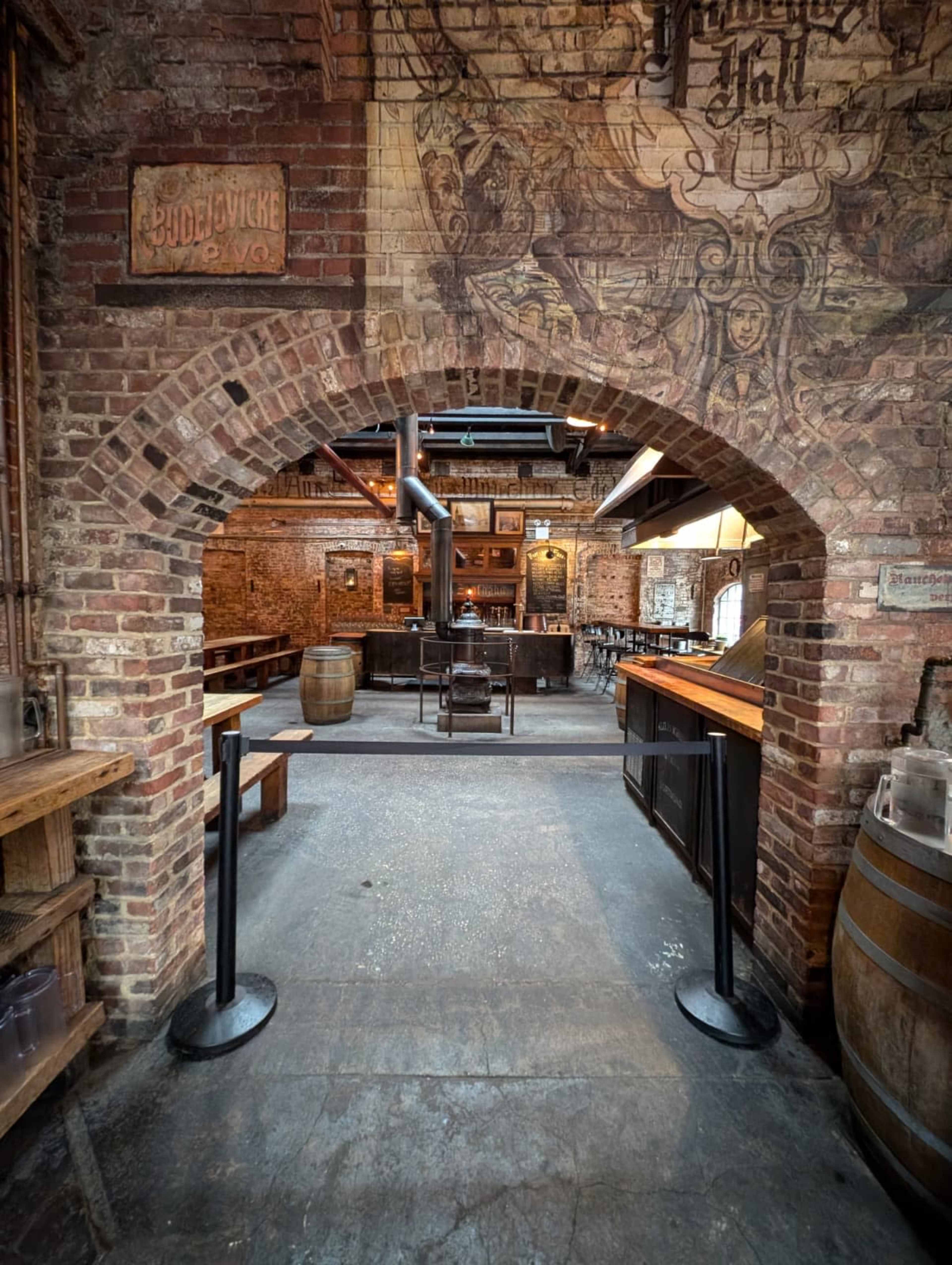 The image shows a rustic interior of a brick-walled bar or restaurant with wooden shelves and a central bar area, illuminated by both natural and artificial light.