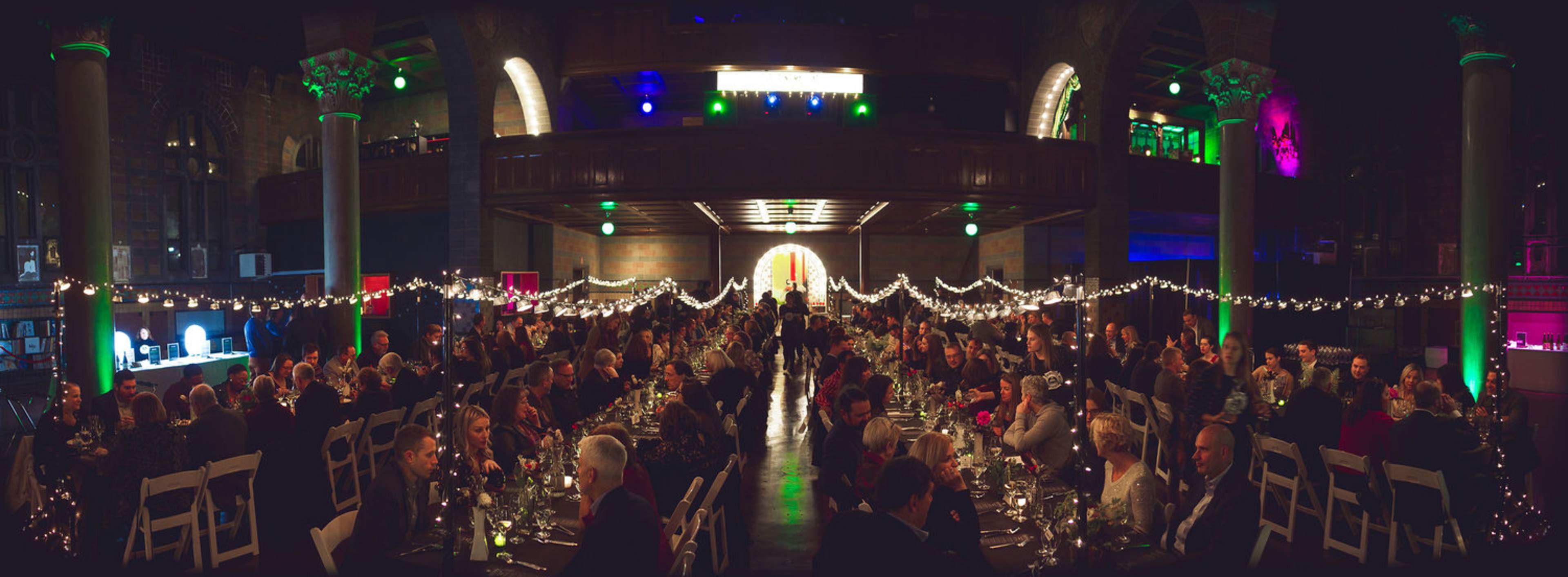The image shows a large indoor event with elegantly set tables filled with guests, adorned with string lights and colorful lighting across a spacious venue.