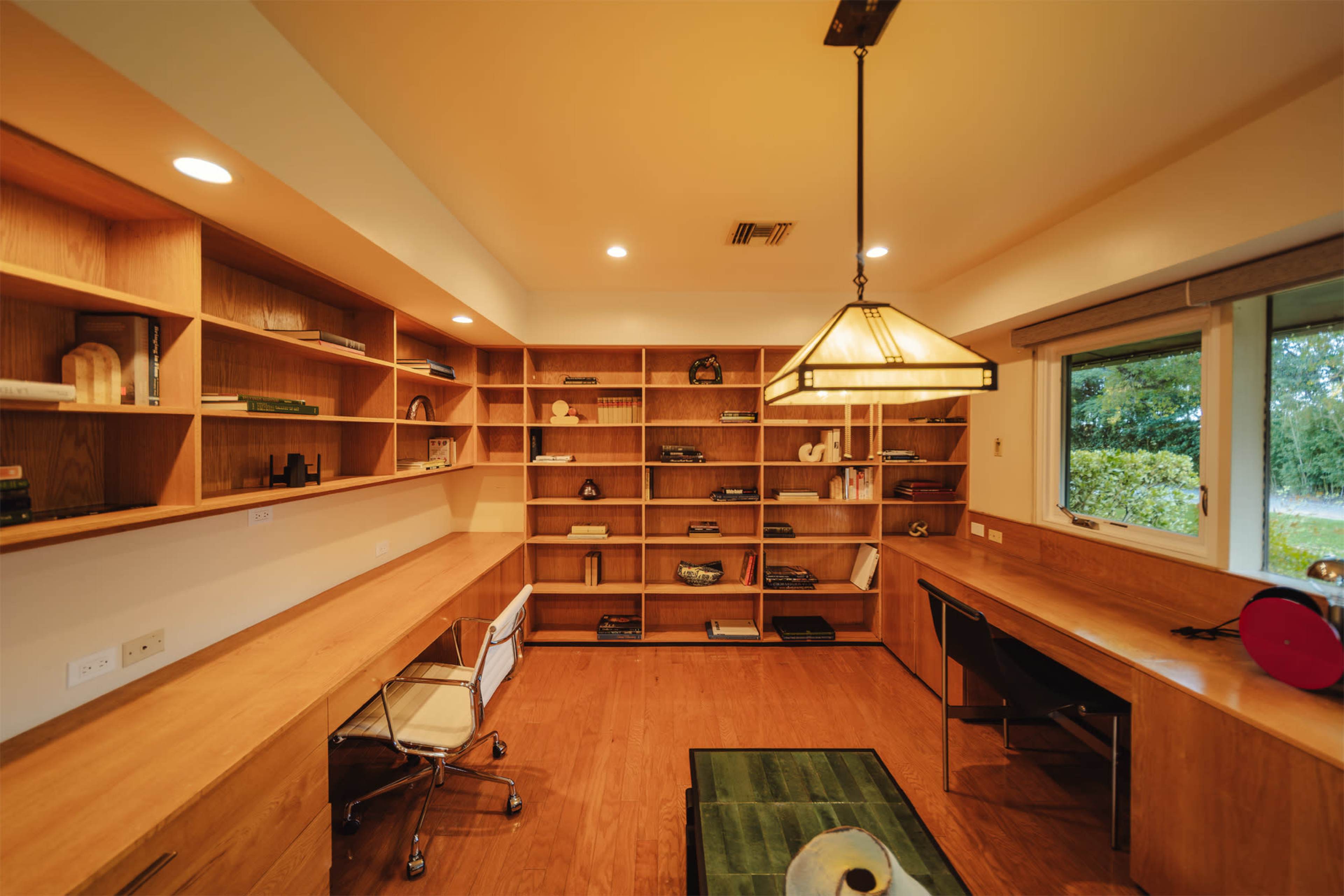 The image shows a spacious, modern home office with wooden shelves, two workstations, and large windows overlooking greenery.