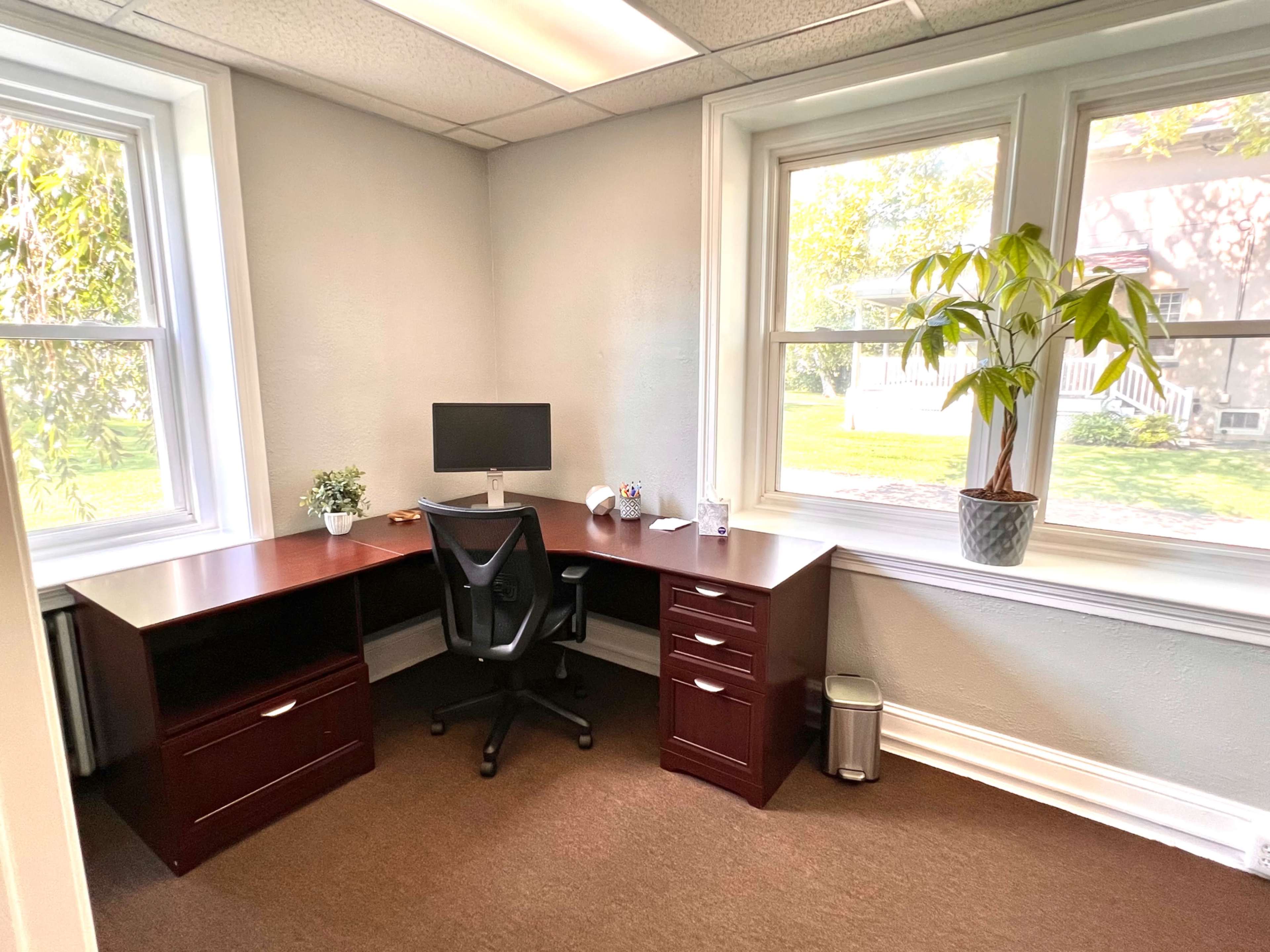 A small office space featuring a wooden desk, an ergonomic chair, a computer monitor, and large windows letting in natural light.