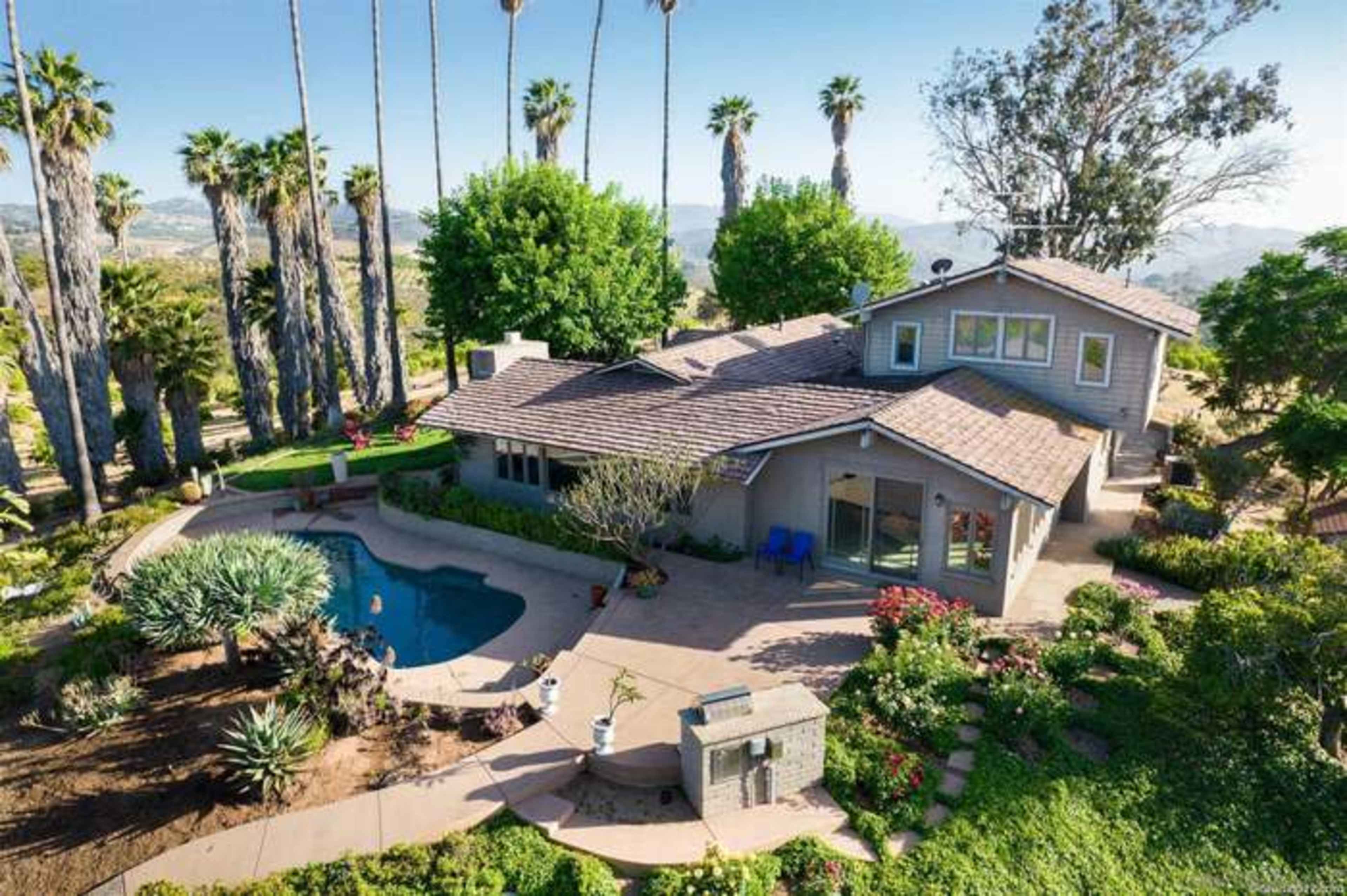 A spacious house with a pool is surrounded by landscaped gardens and tall palm trees, set against a mountainous backdrop.