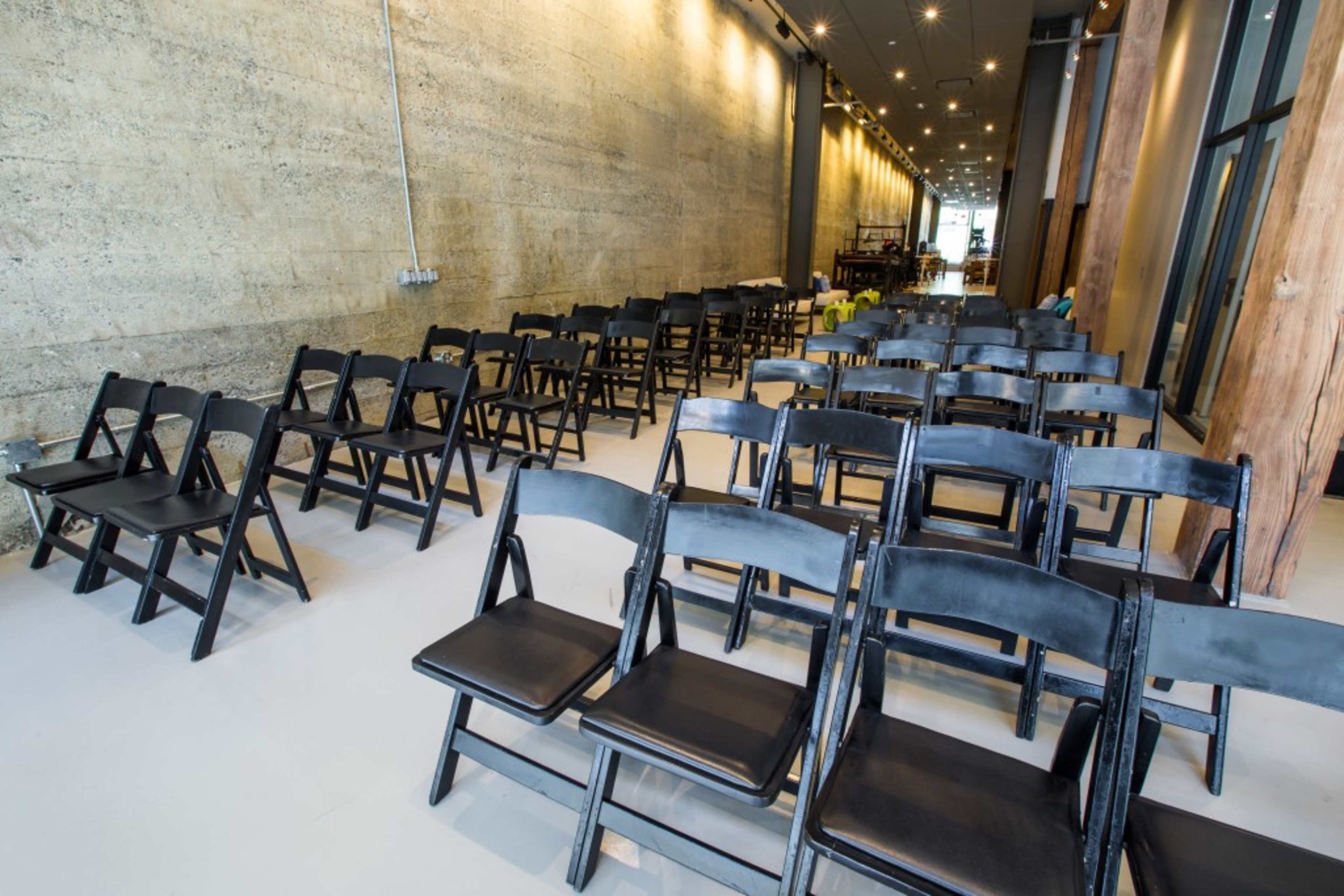 A row of black folding chairs is set up in a spacious interior with a concrete wall and bright lighting.