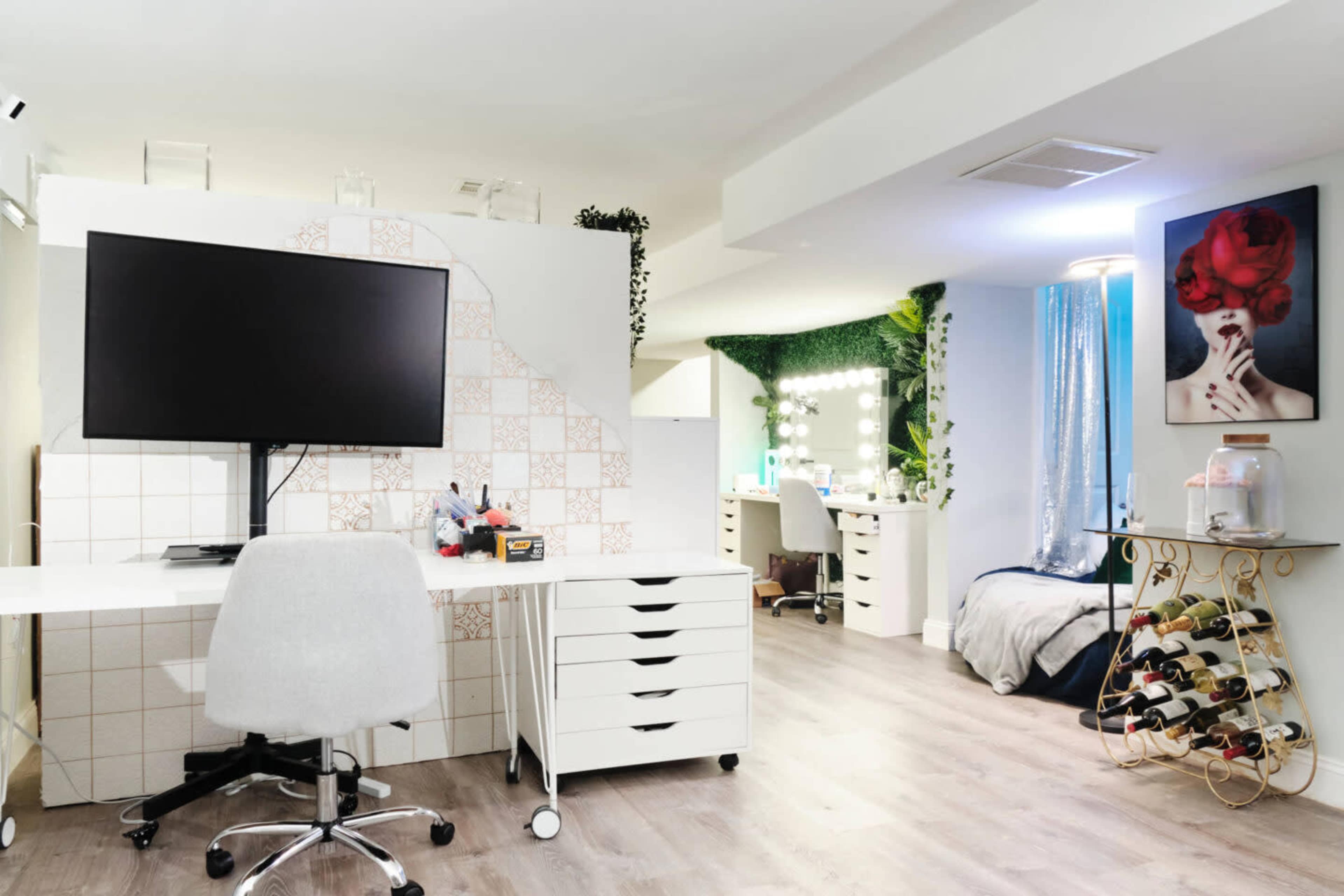A modern home office space featuring a large monitor on a white desk, a chair, storage drawers, and a well-lit vanity area in the background surrounded by greenery.